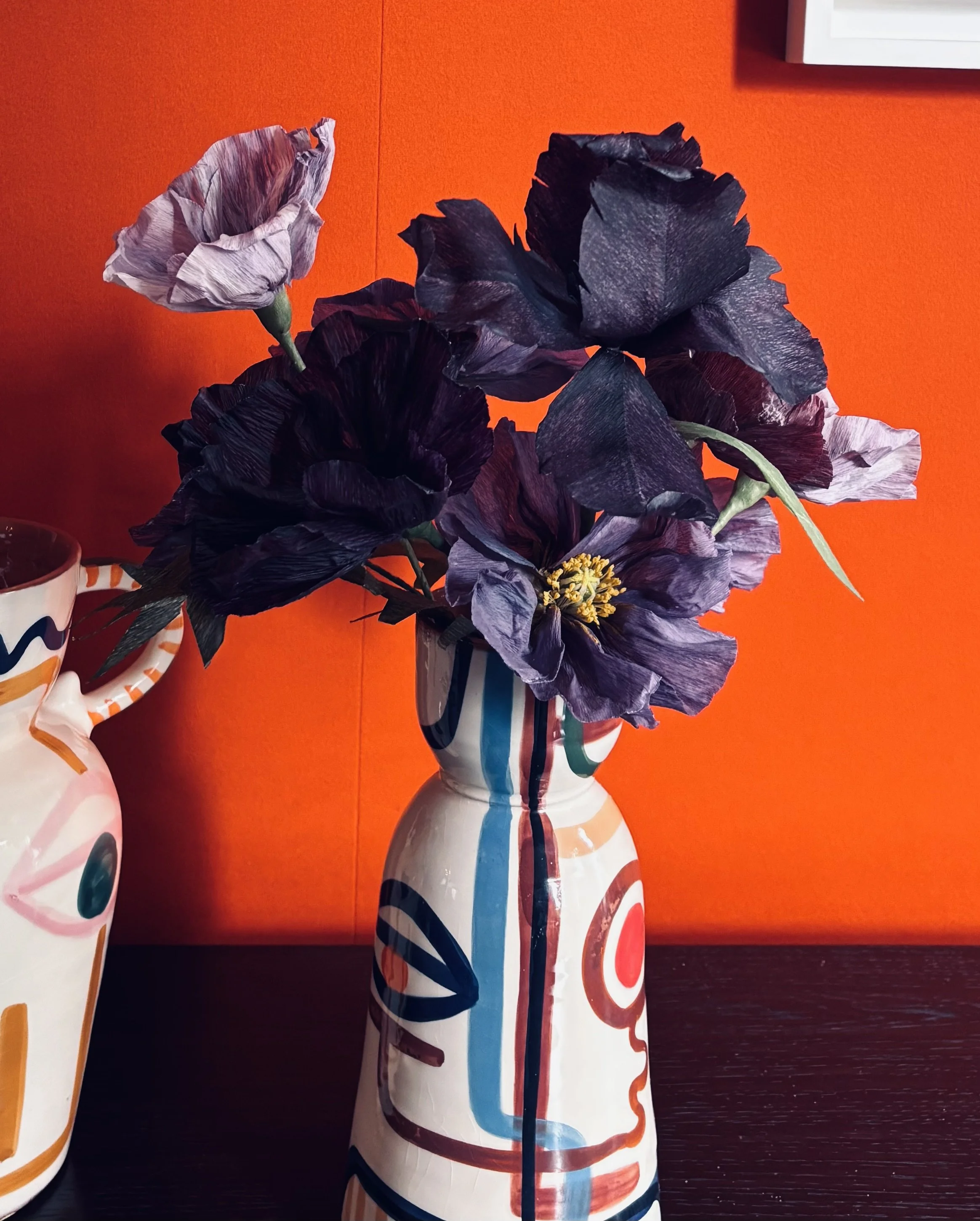Vase with artificial dark purple flowers placed on a table with a colorful abstract tablecloth against an orange background.