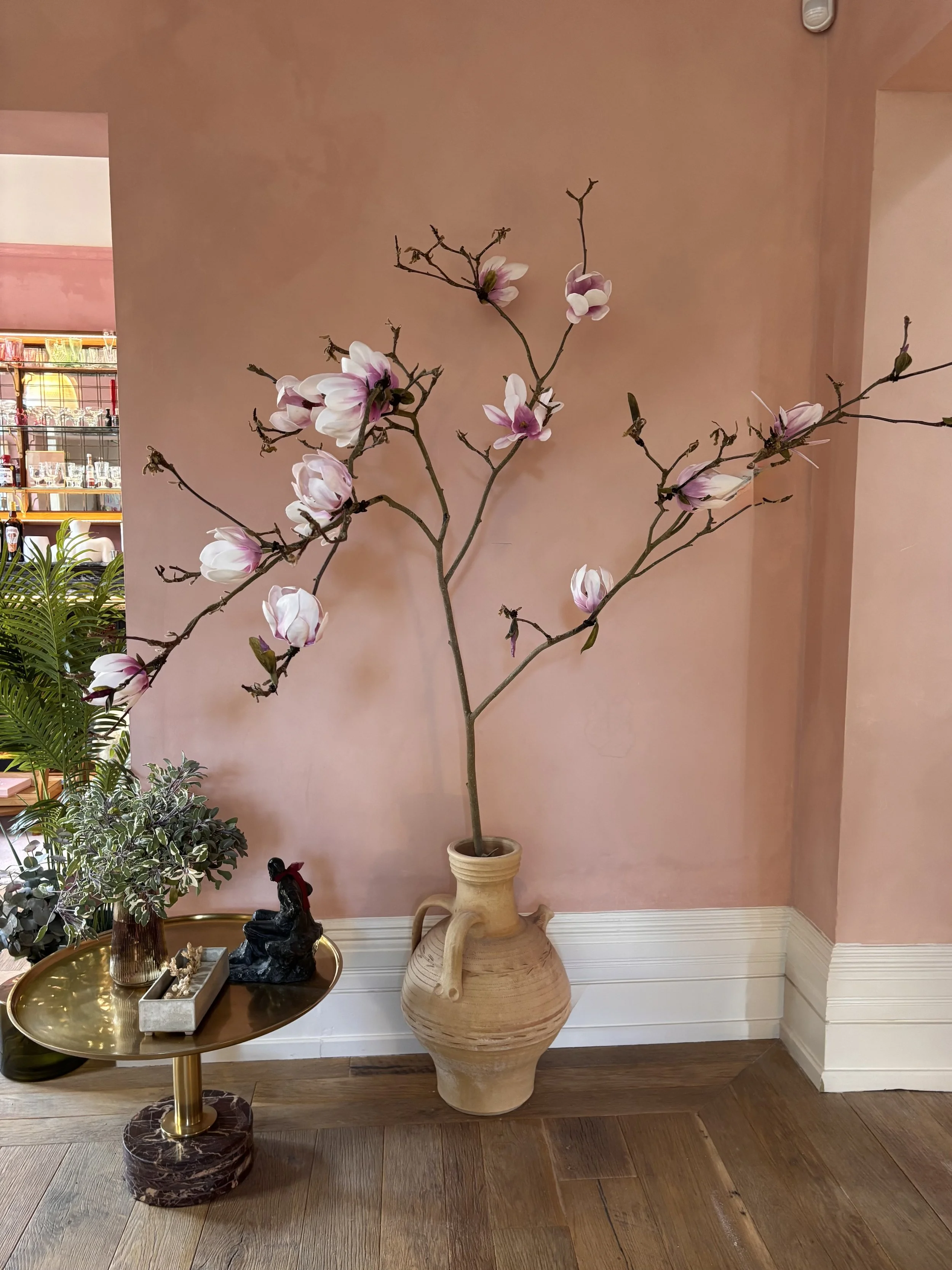 Indoor plant display featuring a large magnolia branch in a large, decorative beige ceramic vase on a wood floor against a pink wall.