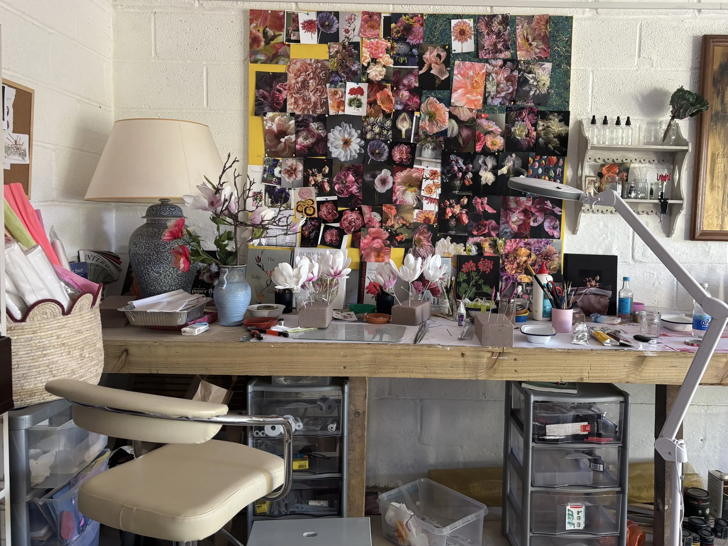 A workspace with a large bulletin board filled with colorful flower photographs, a table with art supplies, vases with flowers, and various office items.