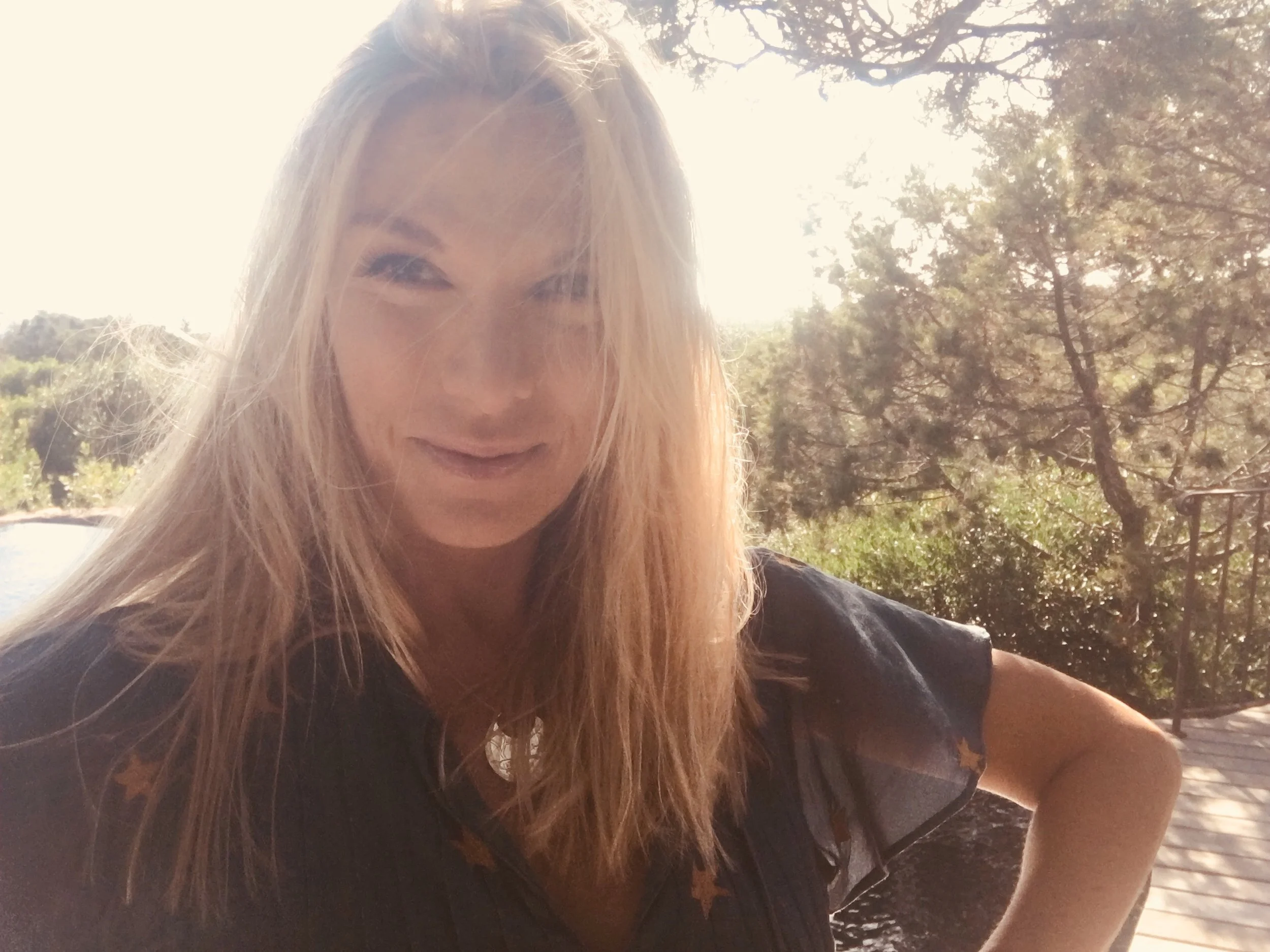 A woman with long blonde hair smiling outdoors with trees in the background, sunlight shining through the trees.