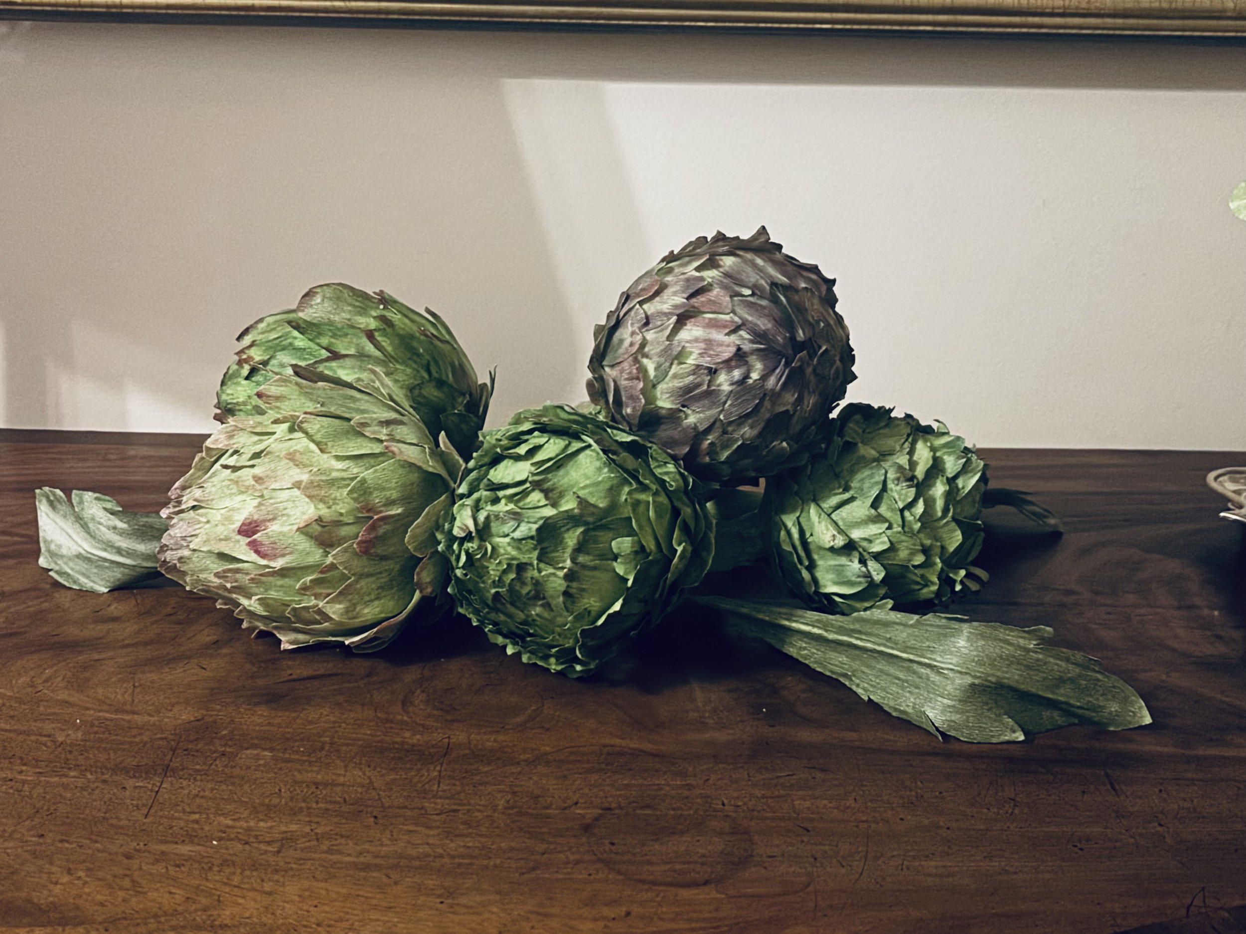 A close-up of artichokes and a leaf on a wooden surface with a white wall background.