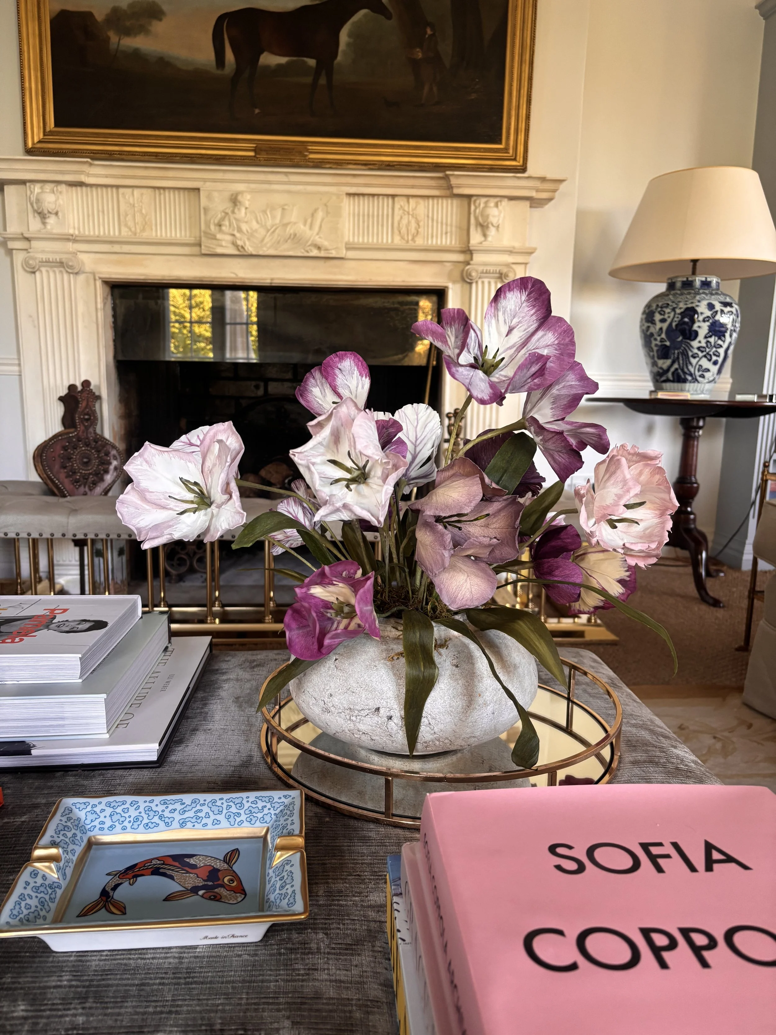 A tabletop with a white vase holding pink and white flowers, surrounded by books and decorative boxes, in a room with a fireplace, a table lamp, and framed artwork.