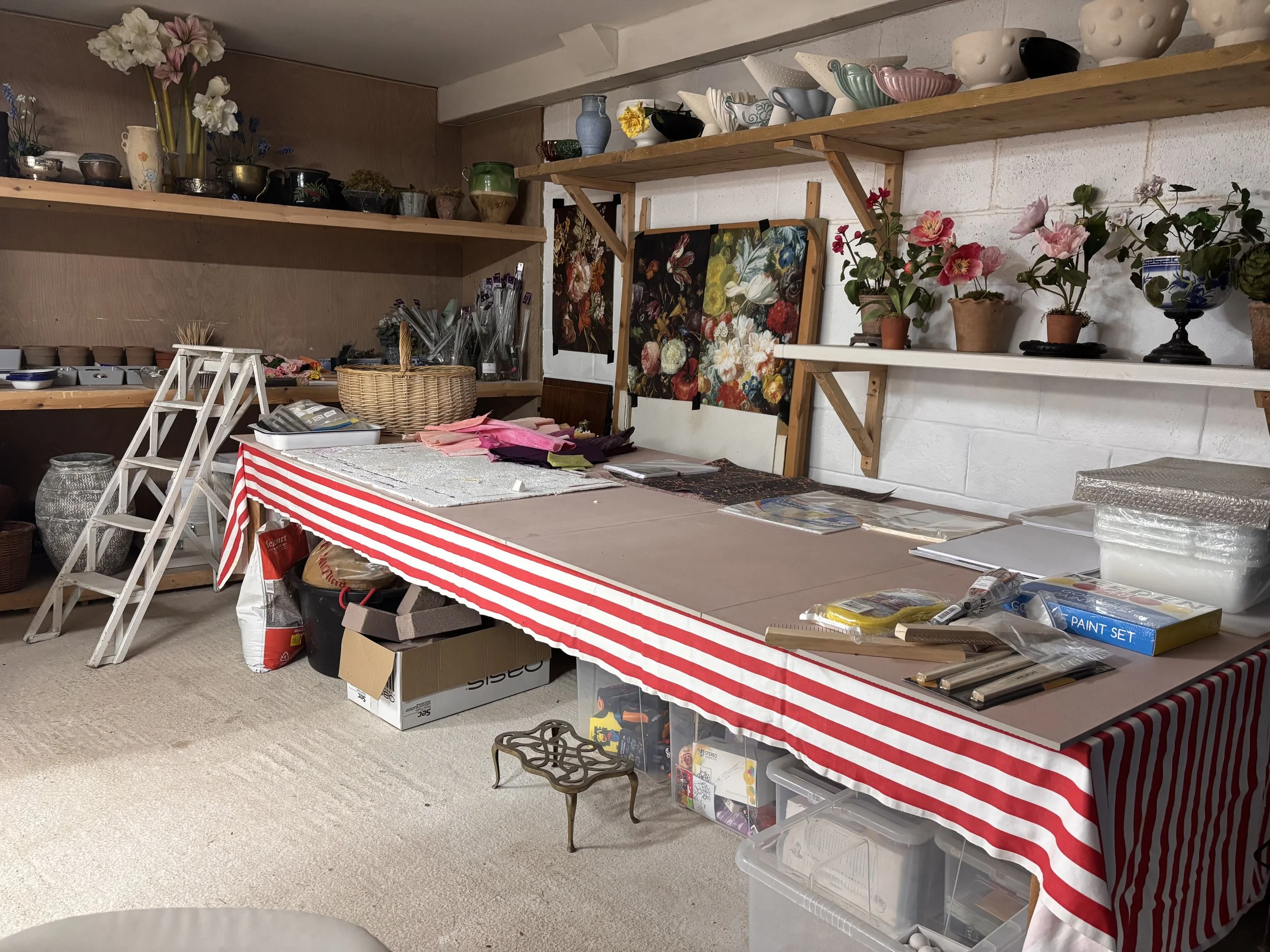 A cluttered craft or resale shop with shelves holding various vases, bowls, and decorative objects, a table covered with supplies including fabric, paper, and tools, and a box labeled 'PAINT SET' under the table.