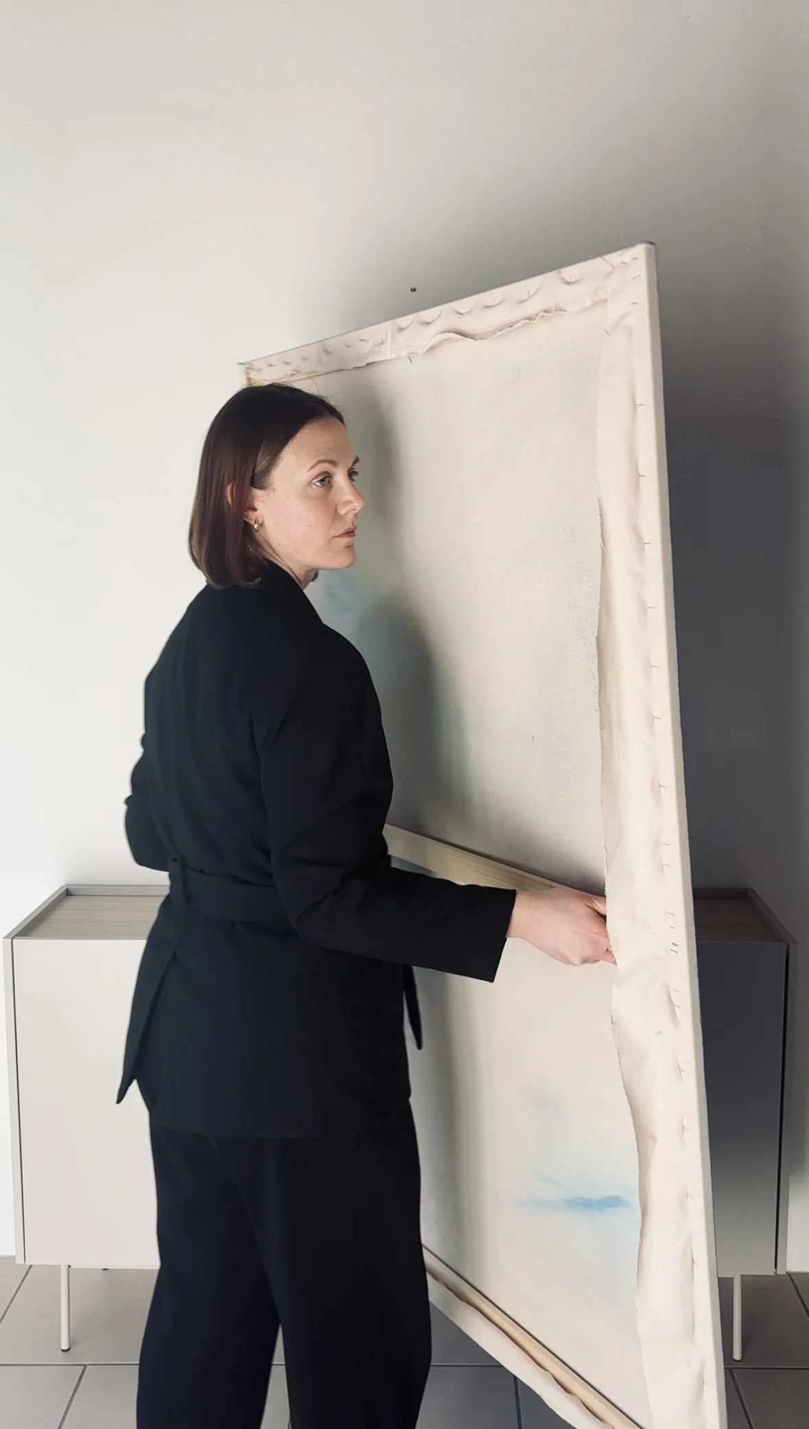 Woman in black clothing standing beside and holding a large blank canvas in a modern room