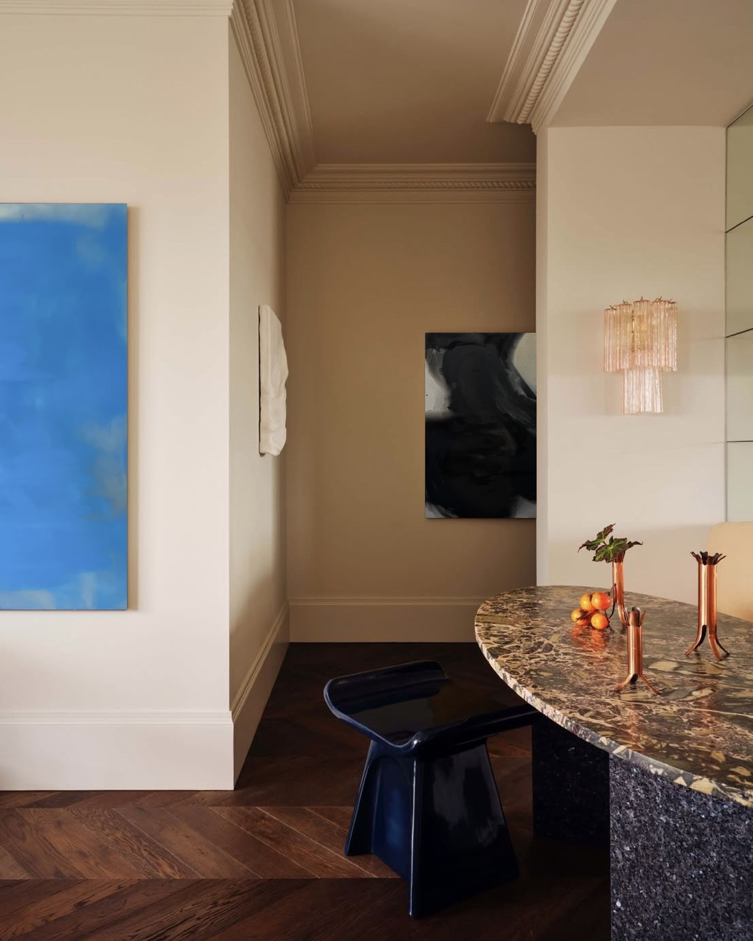 Elegant home interior with blue abstract wall art, ornate molding, black artwork, crystal pendant light, marble table, fruit, and blue chair.