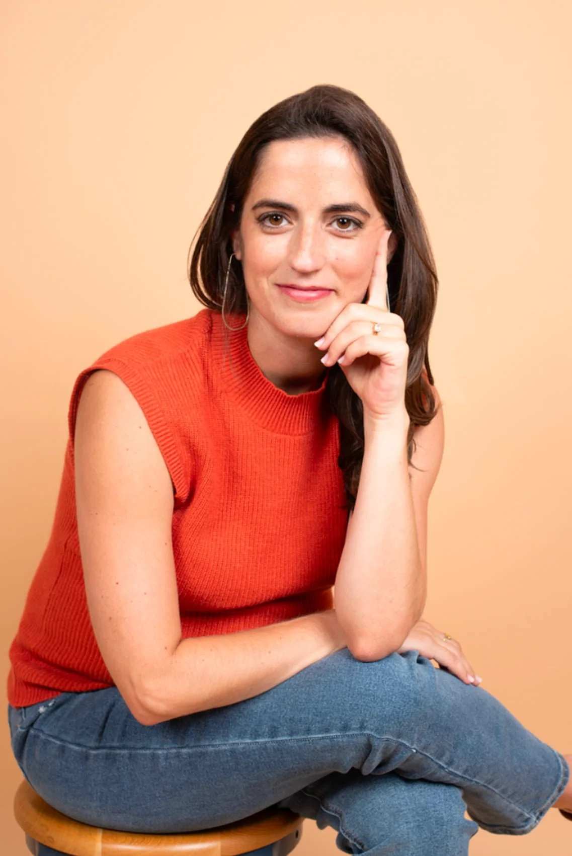 A woman with long brown hair wearing a sleeveless orange top and blue jeans, sitting on a wooden stool against a light beige background, with her right hand resting on her chin.