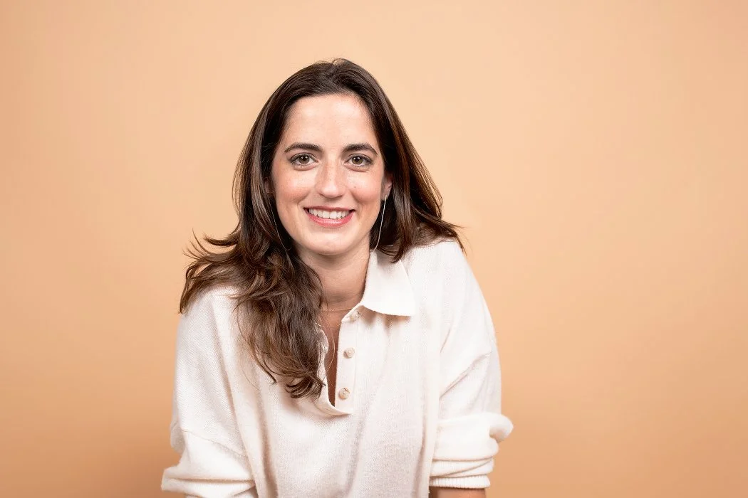 A smiling woman with long brown hair wearing a white shirt, against a beige background.