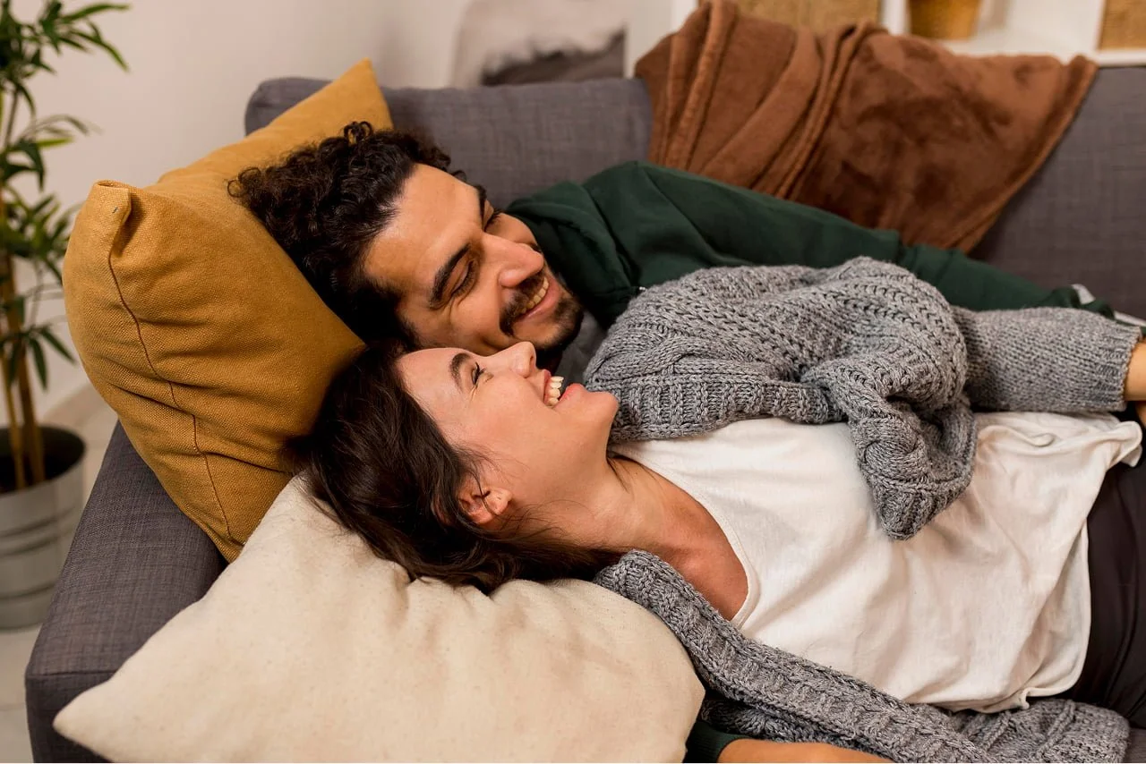 A couple is lying on a couch, smiling and looking at each other. The woman is lying on her back with her head on a pillow, and the man is resting his head close to hers, with one arm around her. They are in a cozy living room with cushions and a potted plant nearby.