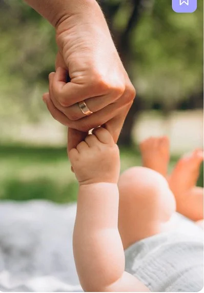 Adult holding baby's small hand outdoors with greenery in the background.