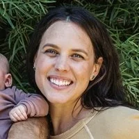 A woman smiling outdoors next to a child, with greenery in the background.