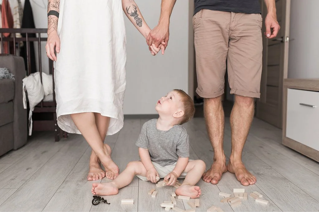 A young boy sitting on the floor, looking up at a woman and man holding hands, with scattered blocks and keys on the floor.