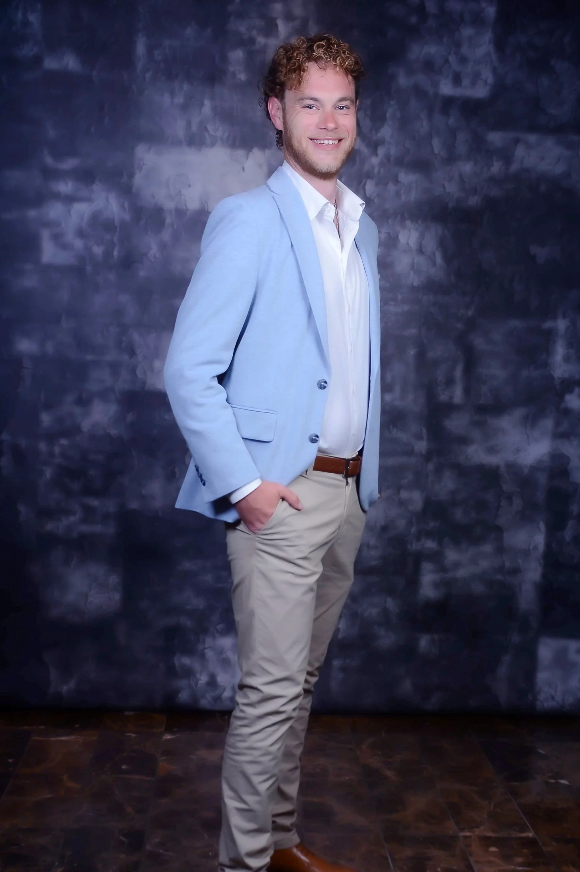A young man with curly hair and a beard, smiling and standing with one hand in his pocket against a dark, textured background. He is wearing a light blue blazer, white shirt, and beige pants.