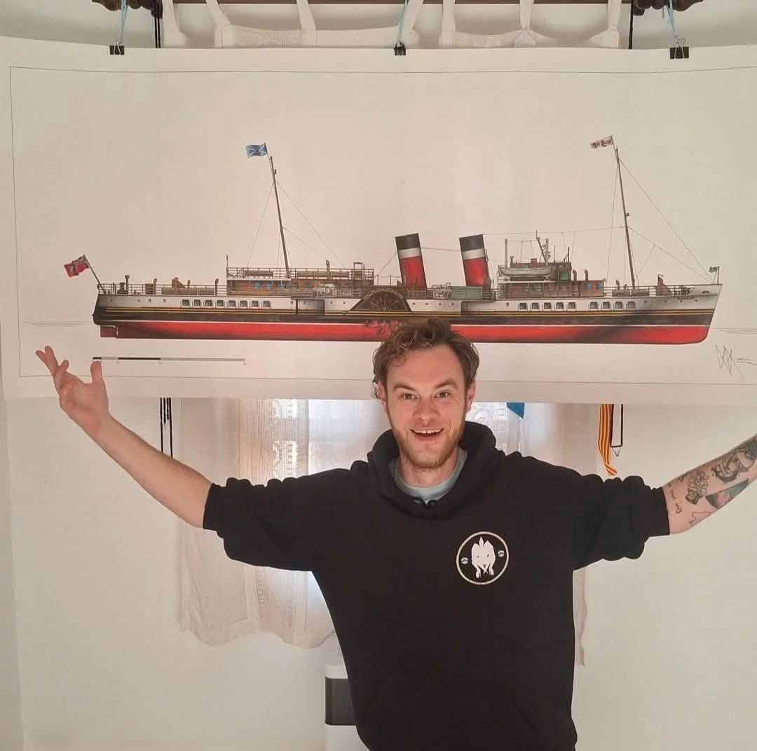 A man with curly hair and tattoos on his right arm smiling and raising his arms, standing in front of a large painting of a vintage ship with red and black smokestacks, a white hull, and flags. Paddlesteamer Waverley