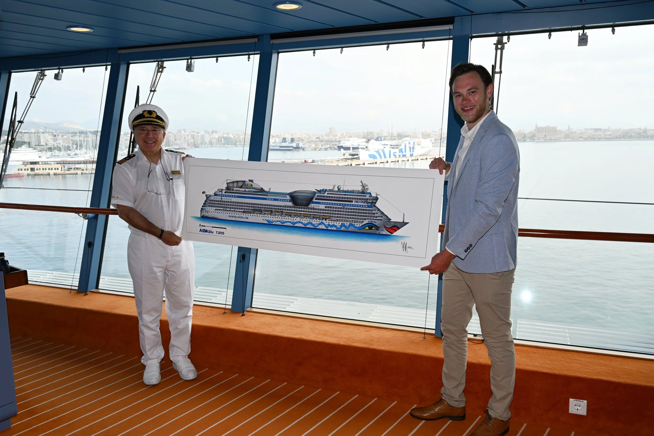 Two men standing inside a boat, holding a large drawing of a cruise ship. One man is in a white uniform with a captain's hat, and the other is in a light gray blazer and beige pants. They are smiling, with a city harbor visible through large windows behind them.