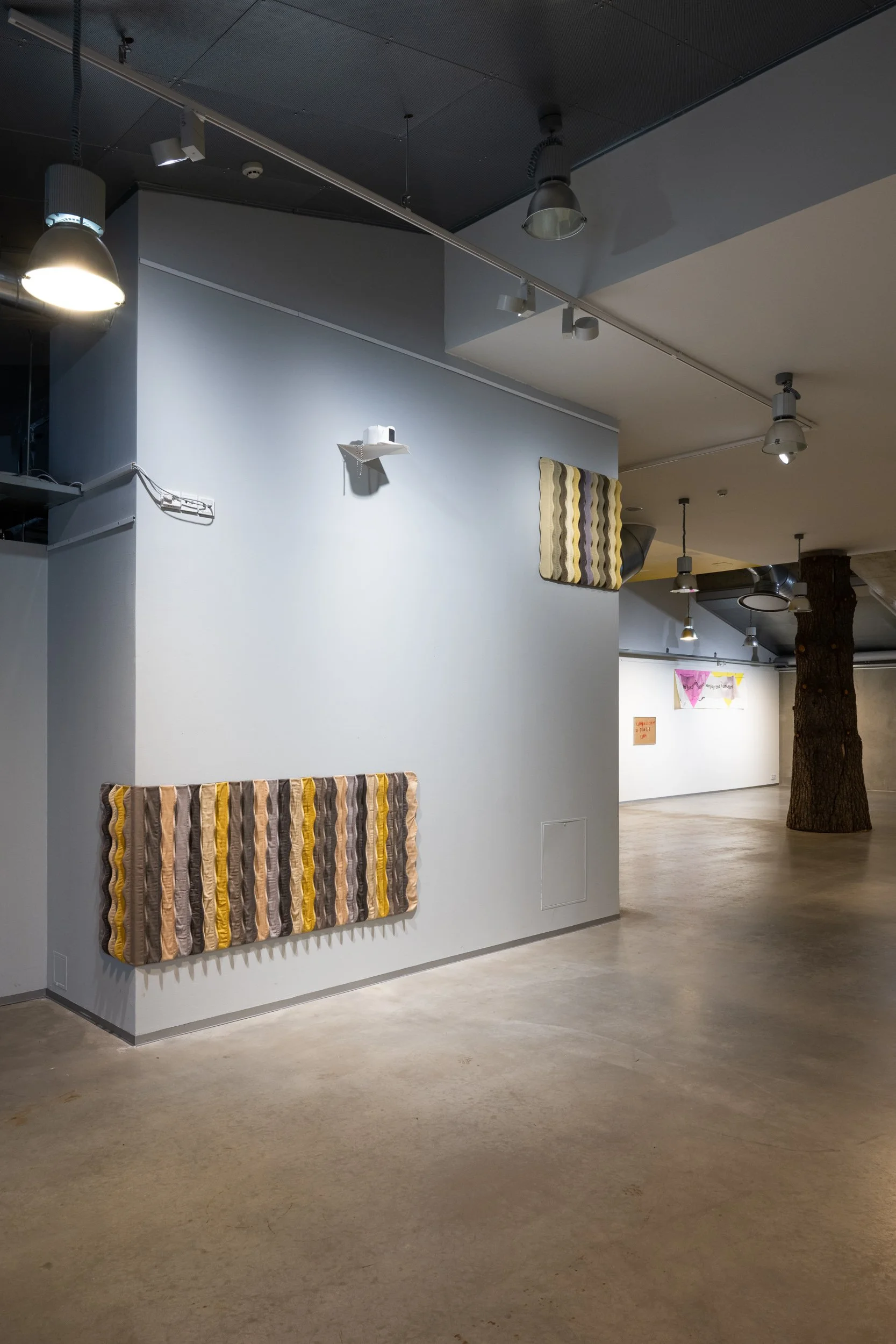 Art gallery interior featuring textured fabric artworks with wavy patterns in earthy tones mounted on a white wall. Modern ceiling lighting and a visible wooden tree trunk in the background.