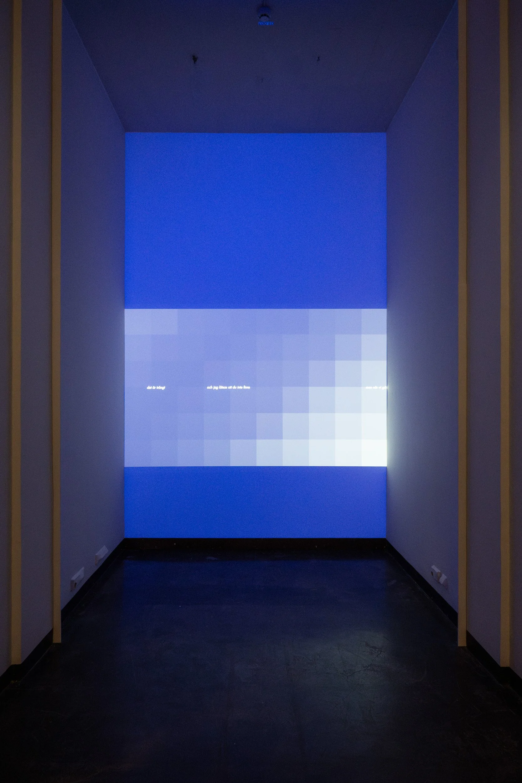 A dark hallway with a large screen at the end displaying a blue pixelated image with some text. The hallway is dimly lit with yellow vertical beams on either side.