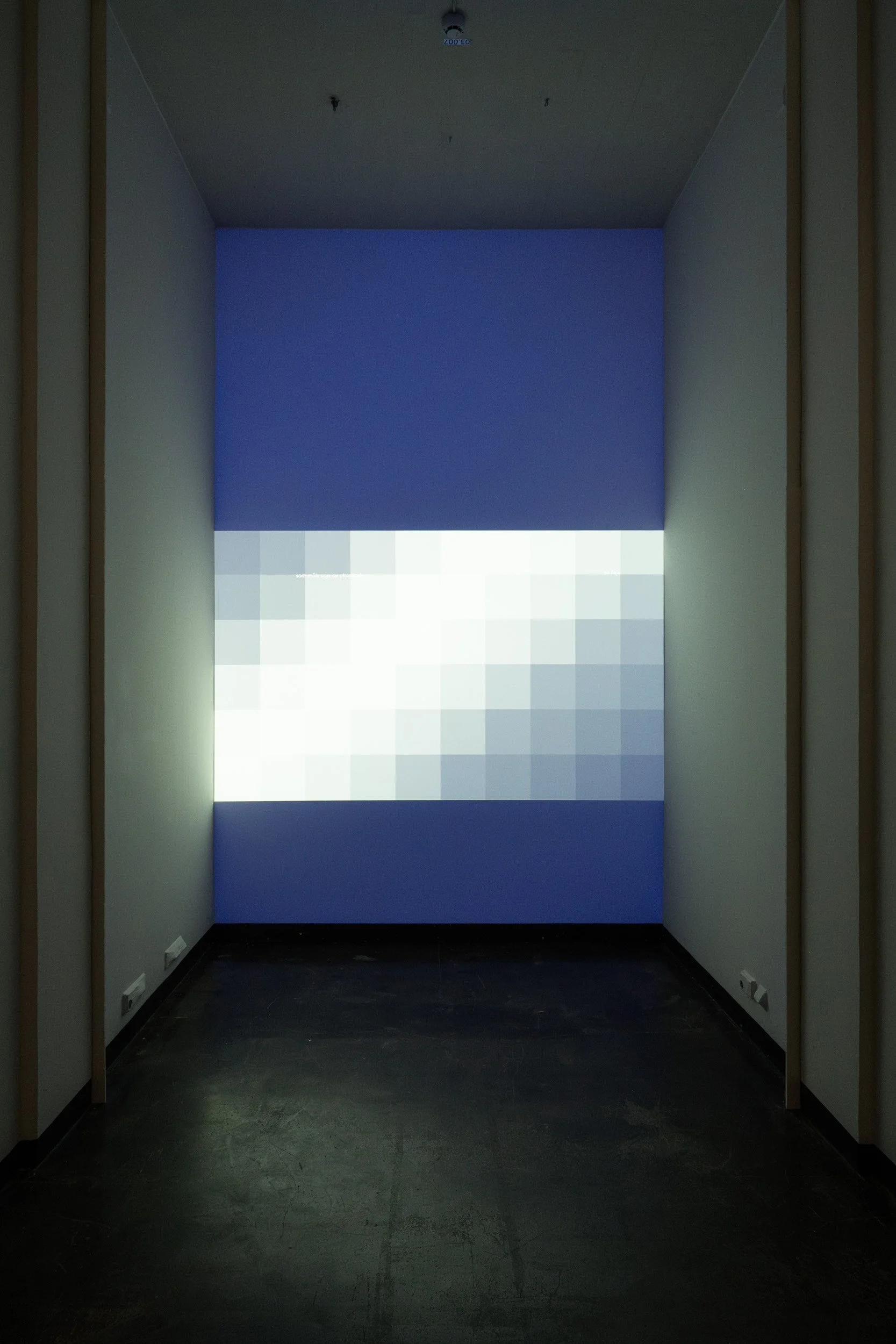 An empty room with a window covered by a blue and white pixelated curtain, dark flooring, and white walls with wooden framing.