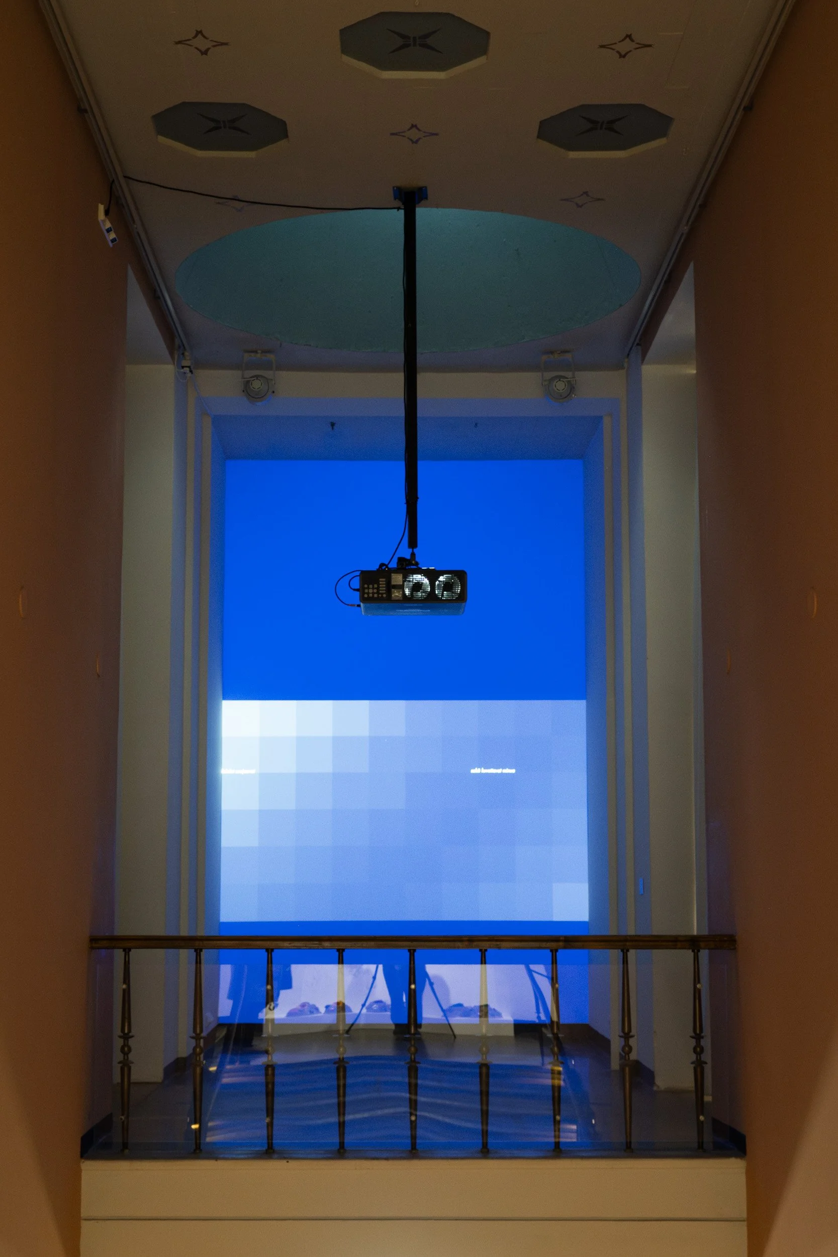 An indoor scene showing a hallway with a balcony and a large projection screen or display at the end. The ceiling has decorative elements, and there is a projector hanging from the ceiling projecting a blue pattern or image onto the screen. The hallw