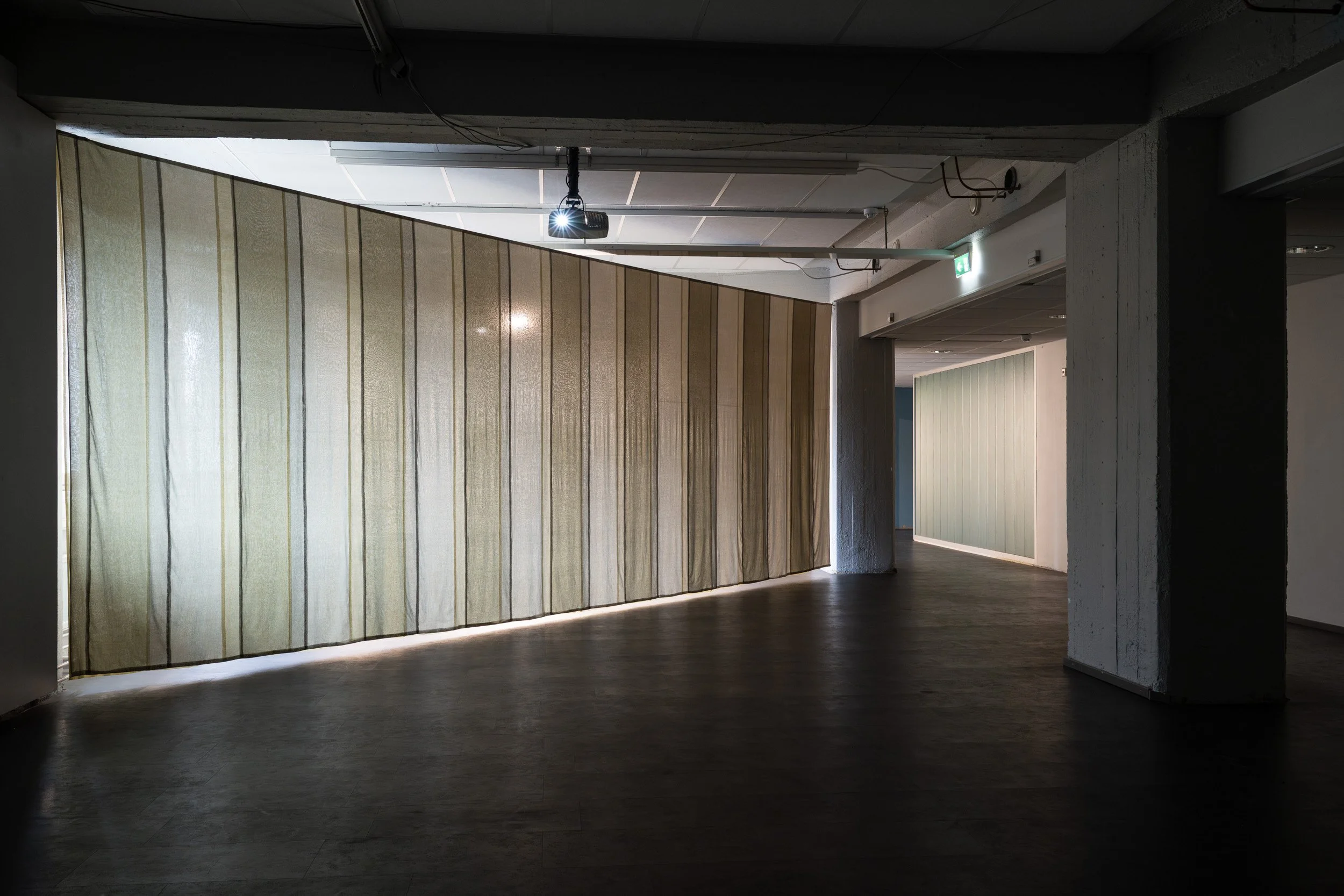 Interior of a building with a large beige curtain hanging from the ceiling, a projector mounted on the ceiling, and a doorway with an illuminated exit sign. The floor appears to be dark wood or laminate.