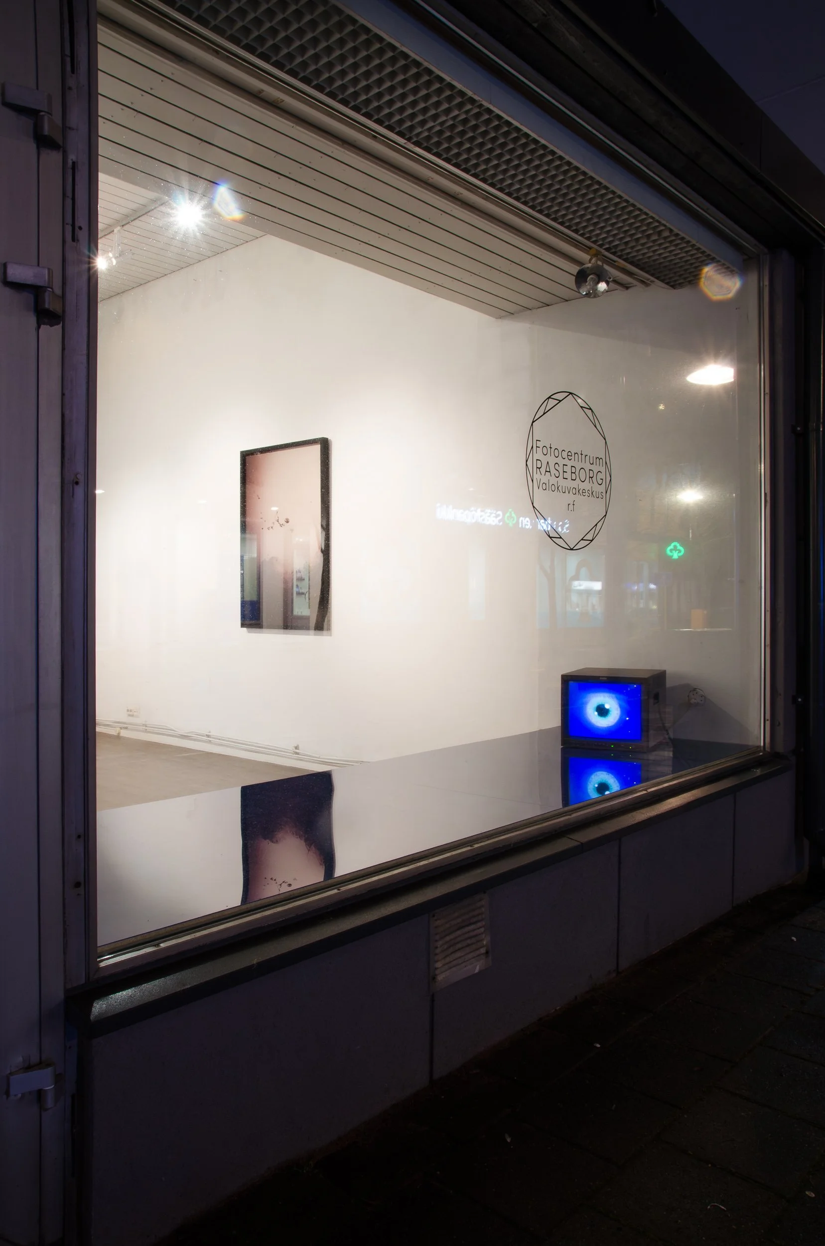 Photo of a gallery window displaying a framed artwork, a sign with 'Fotocentrum RASEBORG Valokuvakeskus,' and a small TV showing a blue eye, with a reflection of a person's face on the glass.