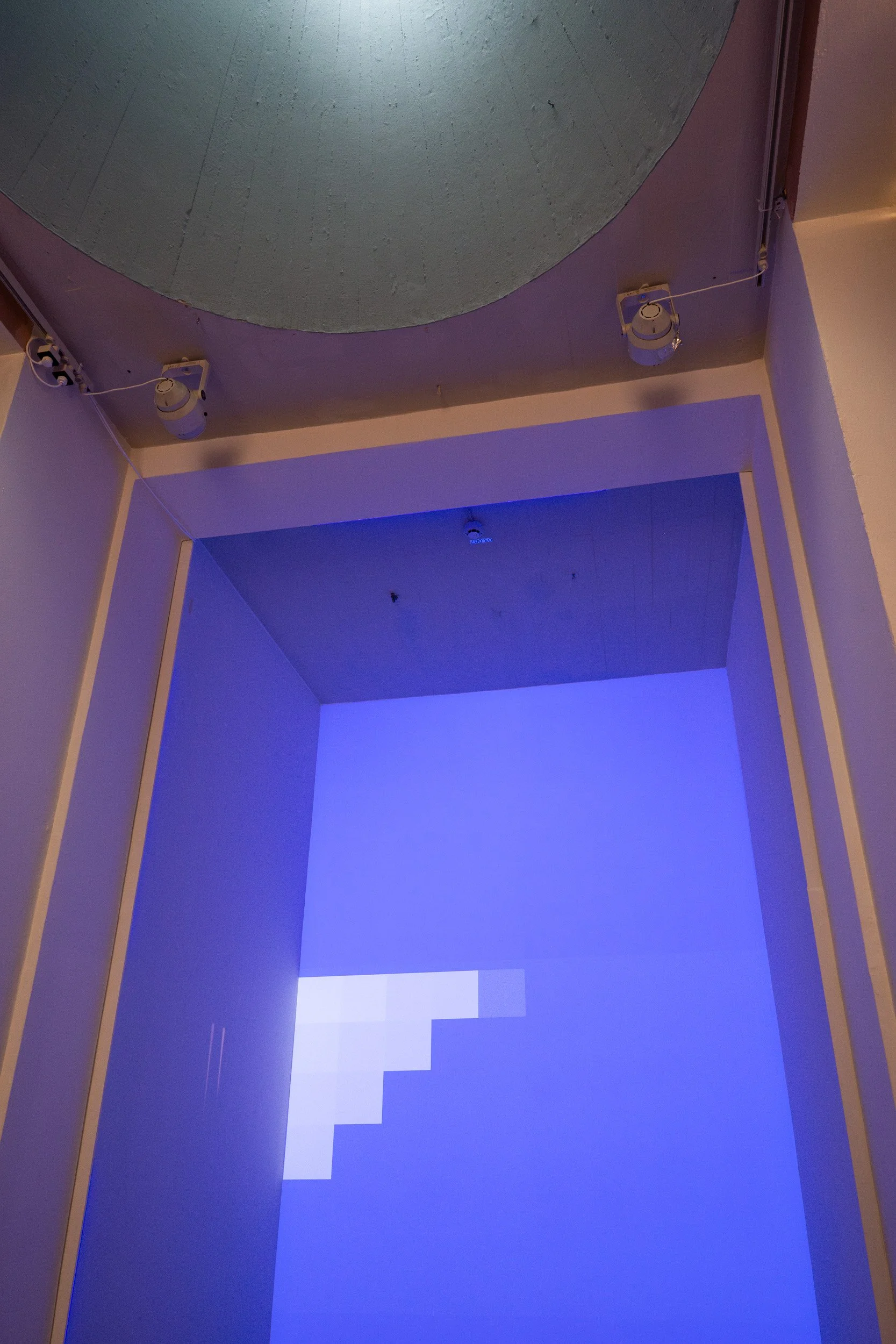 A hallway with purple and beige walls and a stepped blue light projection on the floor. The ceiling has spotlights and a circular design element.