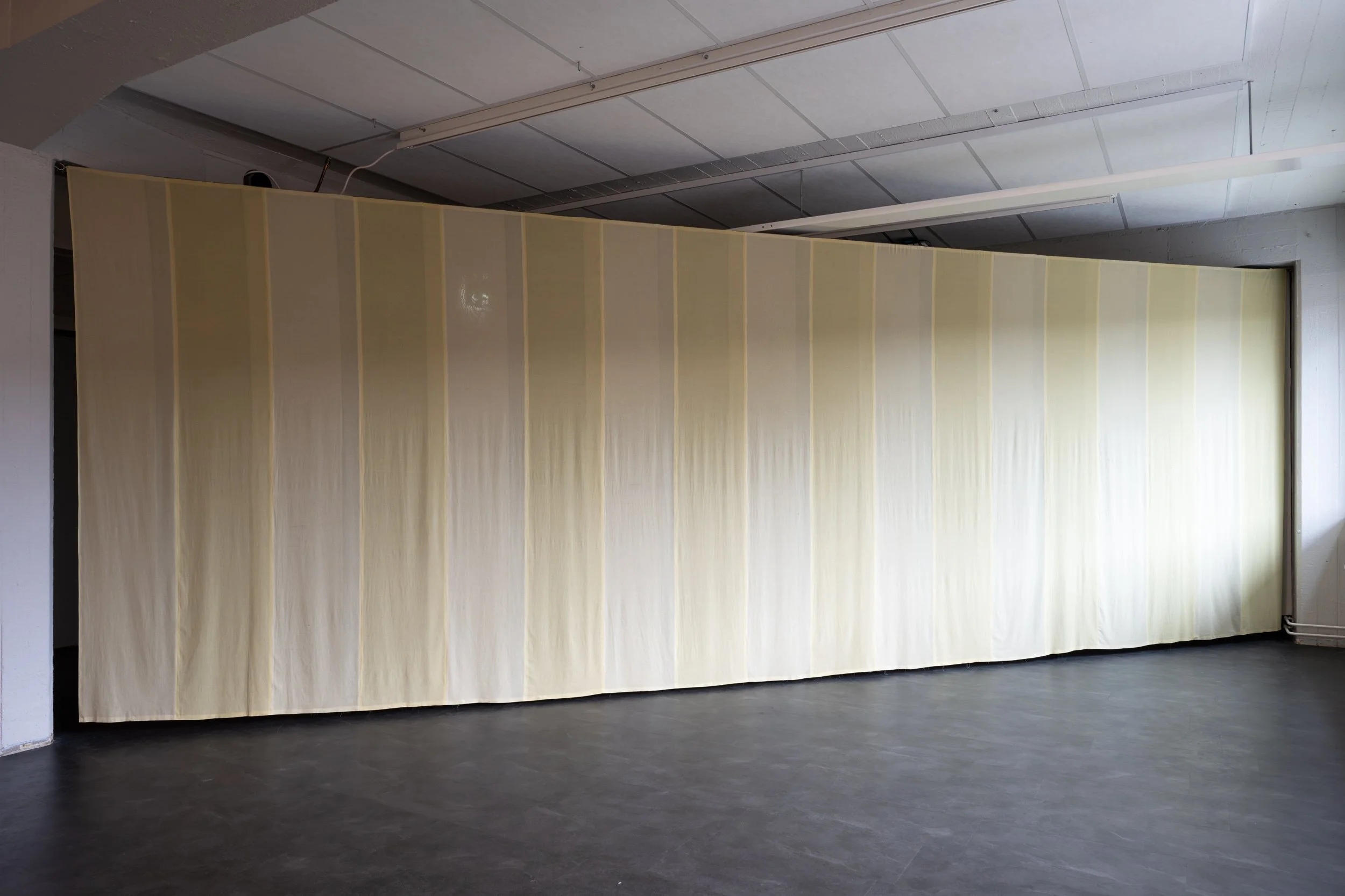 An indoor space with a large, beige partition or wall and a dark gray floor. The ceiling has visible white panels and some lighting fixtures. There's a window or opening on the right side of the wall.