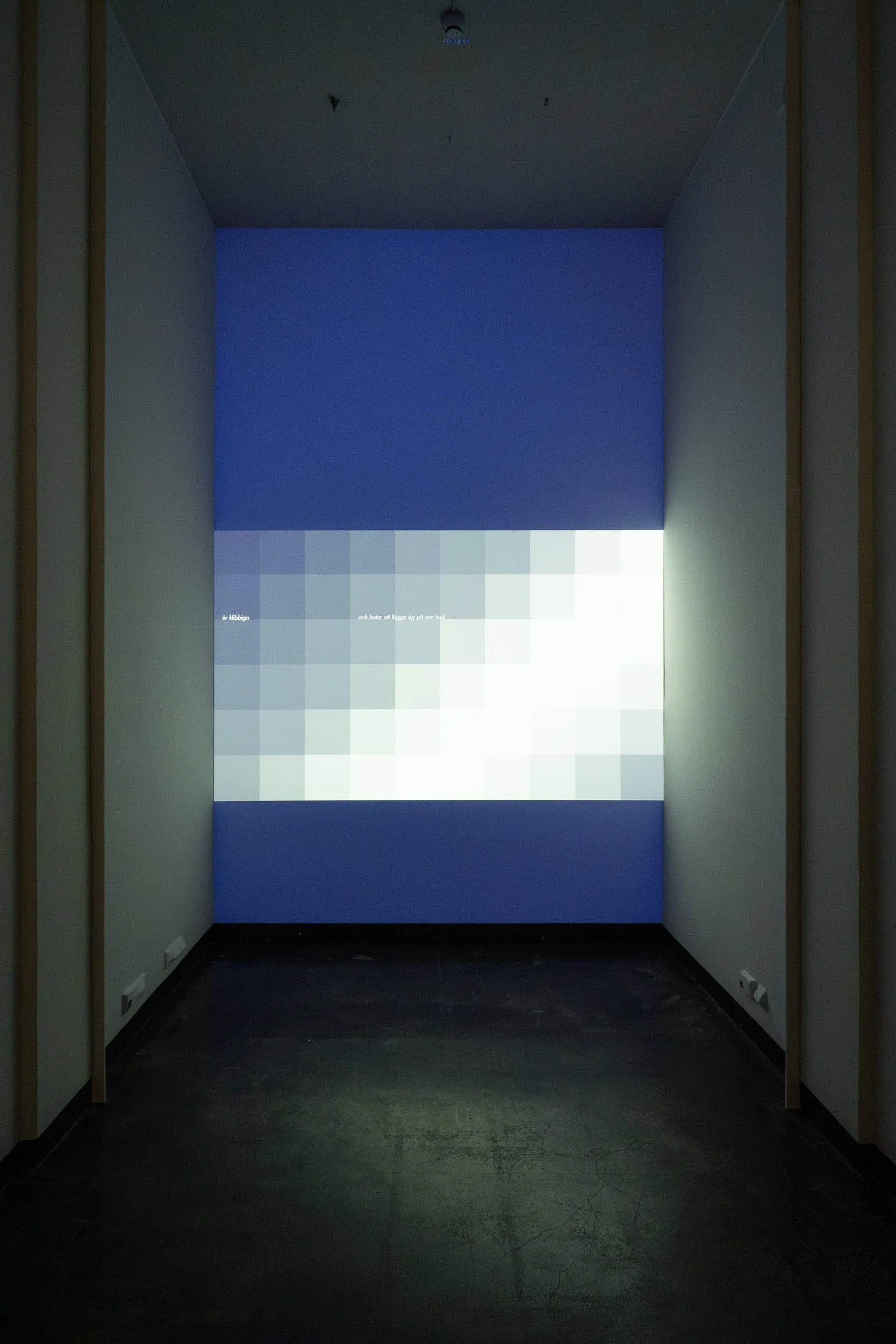 Exhibit space with a bright digital screen on the back wall displaying pixelated blue and white graphics and text, in a dark room with minimal lighting and black flooring.