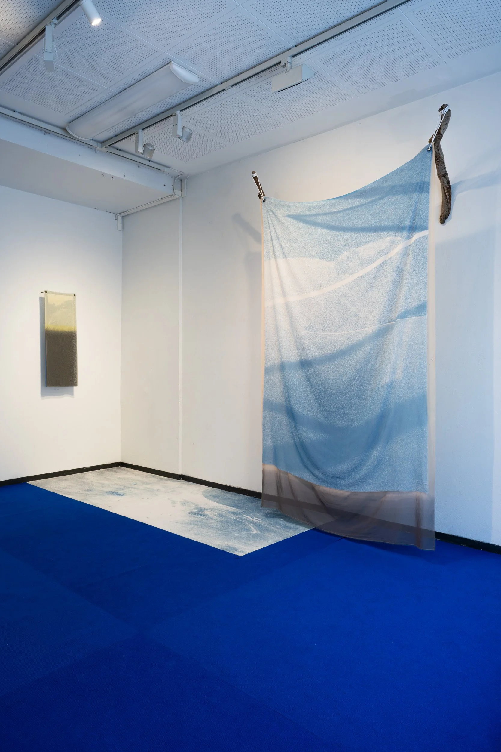 An abstract art installation in a gallery with a blue floor, a white wall, a fabric panel hanging from the ceiling, a rectangular wall piece, and a painted area on the floor.