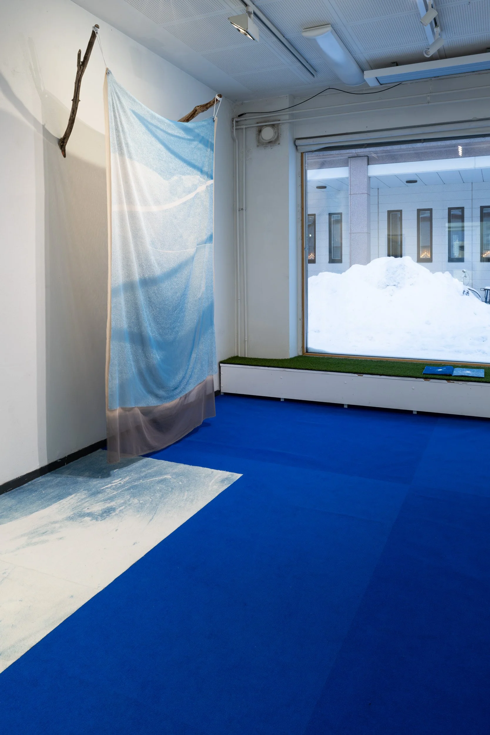 Interior of a room with blue and white flooring, a large window showing a snowy scene outside, and a fabric display with tree branches as support.