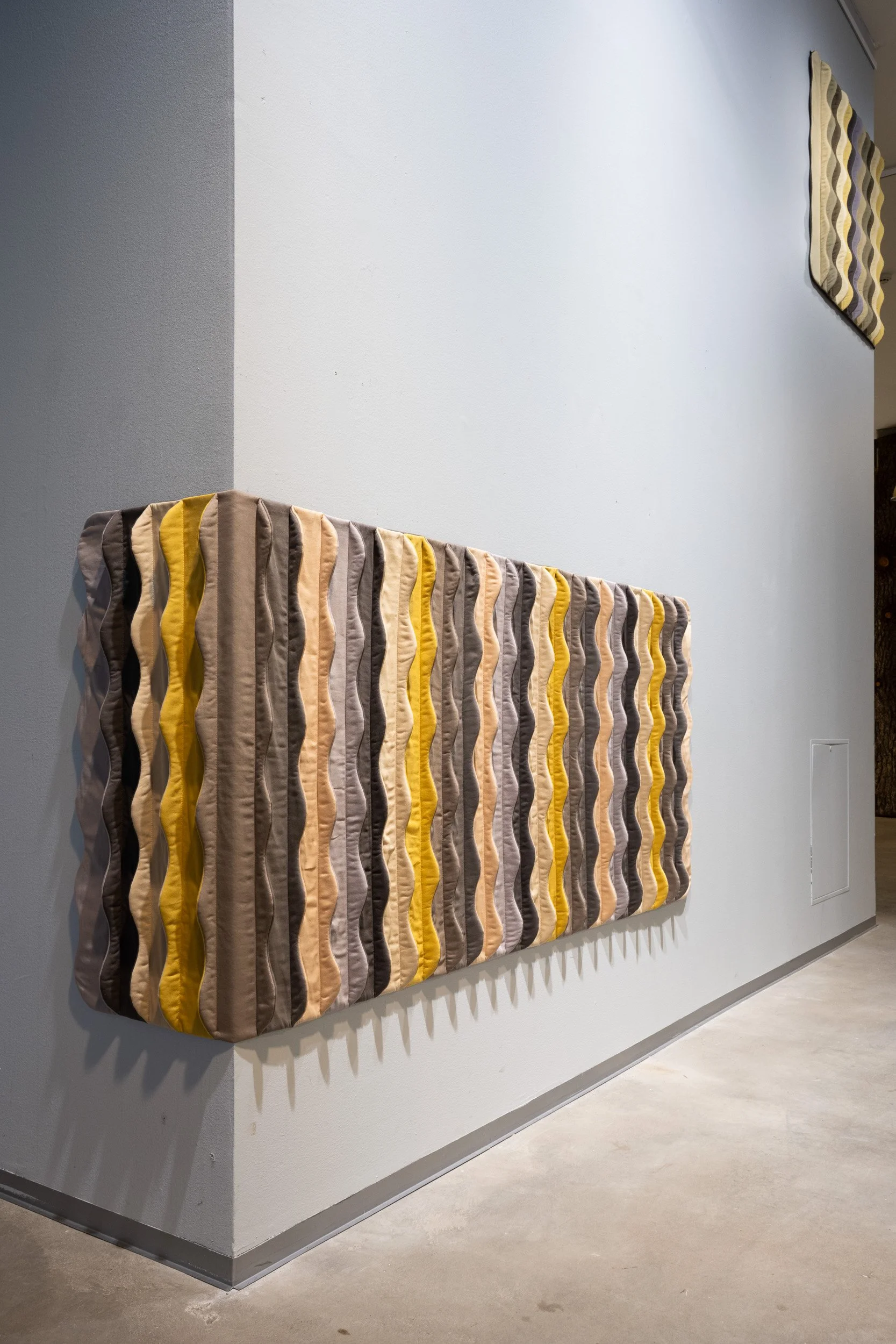 Wall art installation featuring a large panel of vertically oriented wavy fabric strips in shades of gray, beige, yellow, and brown, mounted on a white wall in a gallery space with a gray floor.