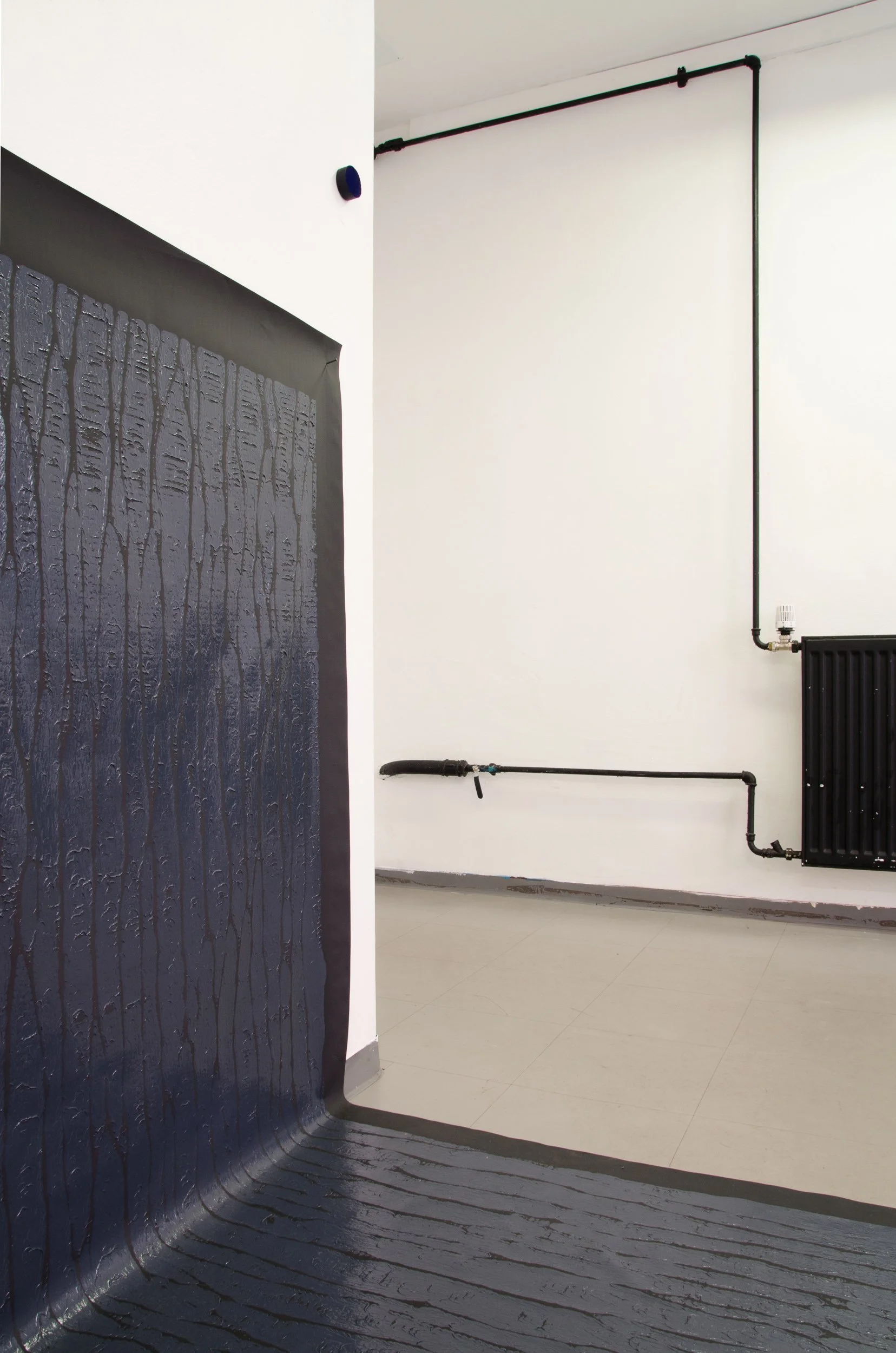 Interior space with a black textured wall on the left and an off-white wall on the right. The off-white wall has black pipes running horizontally and vertically, connected to a radiator. The floor is white tiled.
