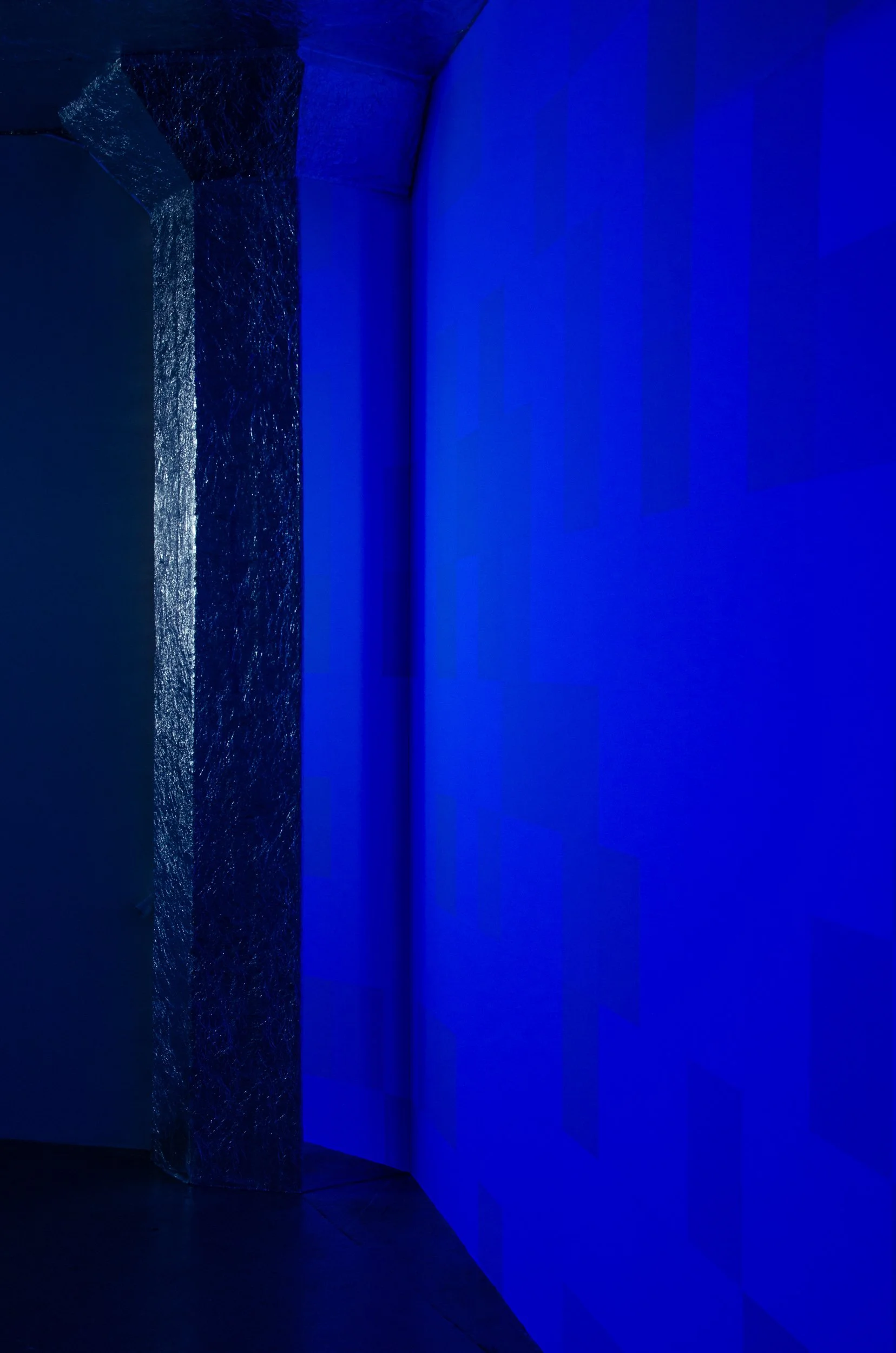 Installation art piece with a large, textured, reflective vertical sculpture illuminated in blue light, placed against a blue wall with projected blue lighting.