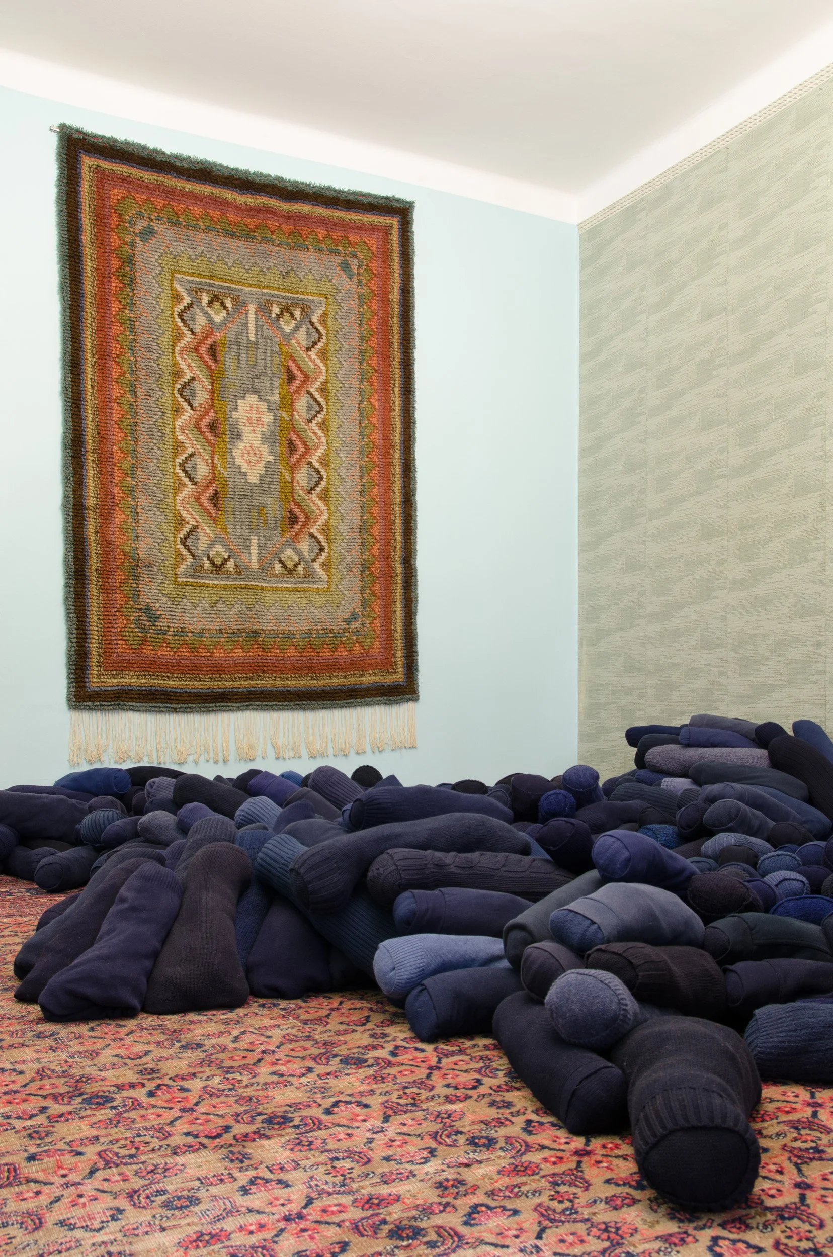 A group of people in black or dark clothing praying on a patterned rug in a room with a pale blue wall, a colorful hanging tapestry, and a light-colored wall. People are lying face down with their arms extended forward.