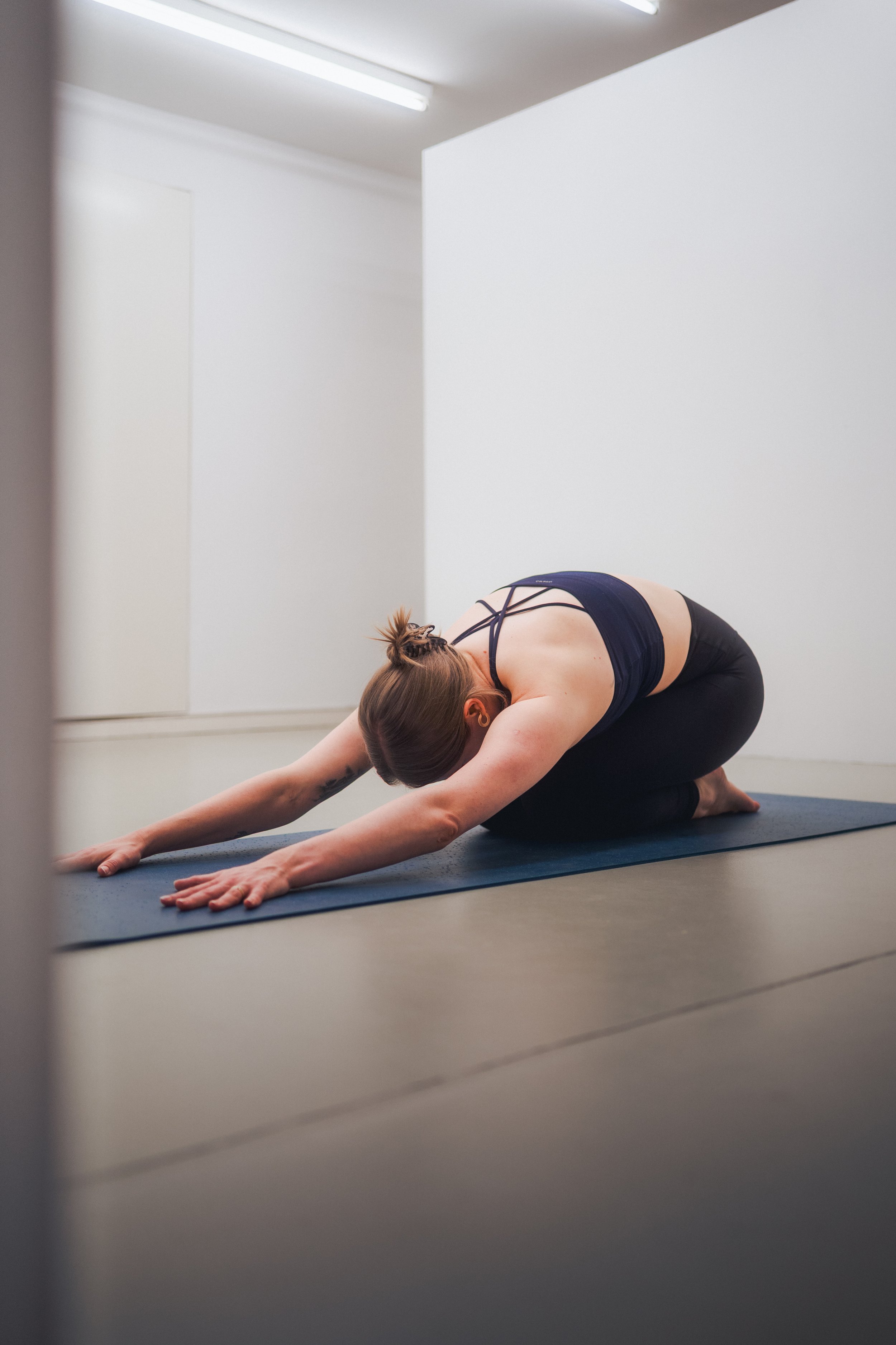 Woman in black athletic clothing practicing child's pose on a yoga mat in a bright studio