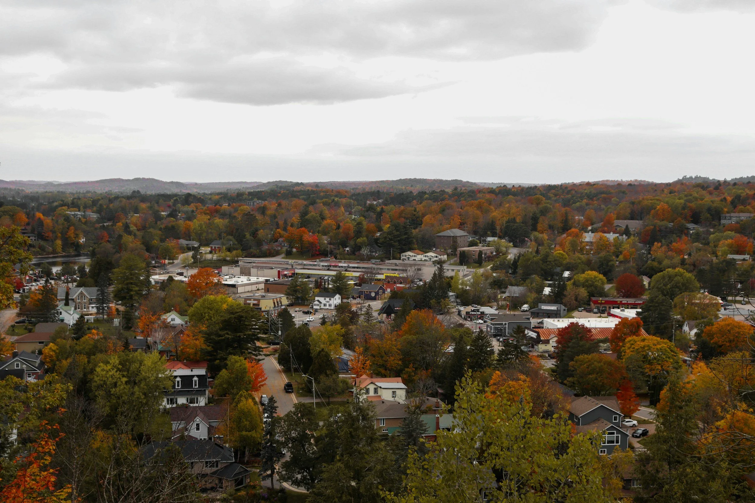 A view of a town surrounded by trees with fall foliage, with a cloudy sky overhead.