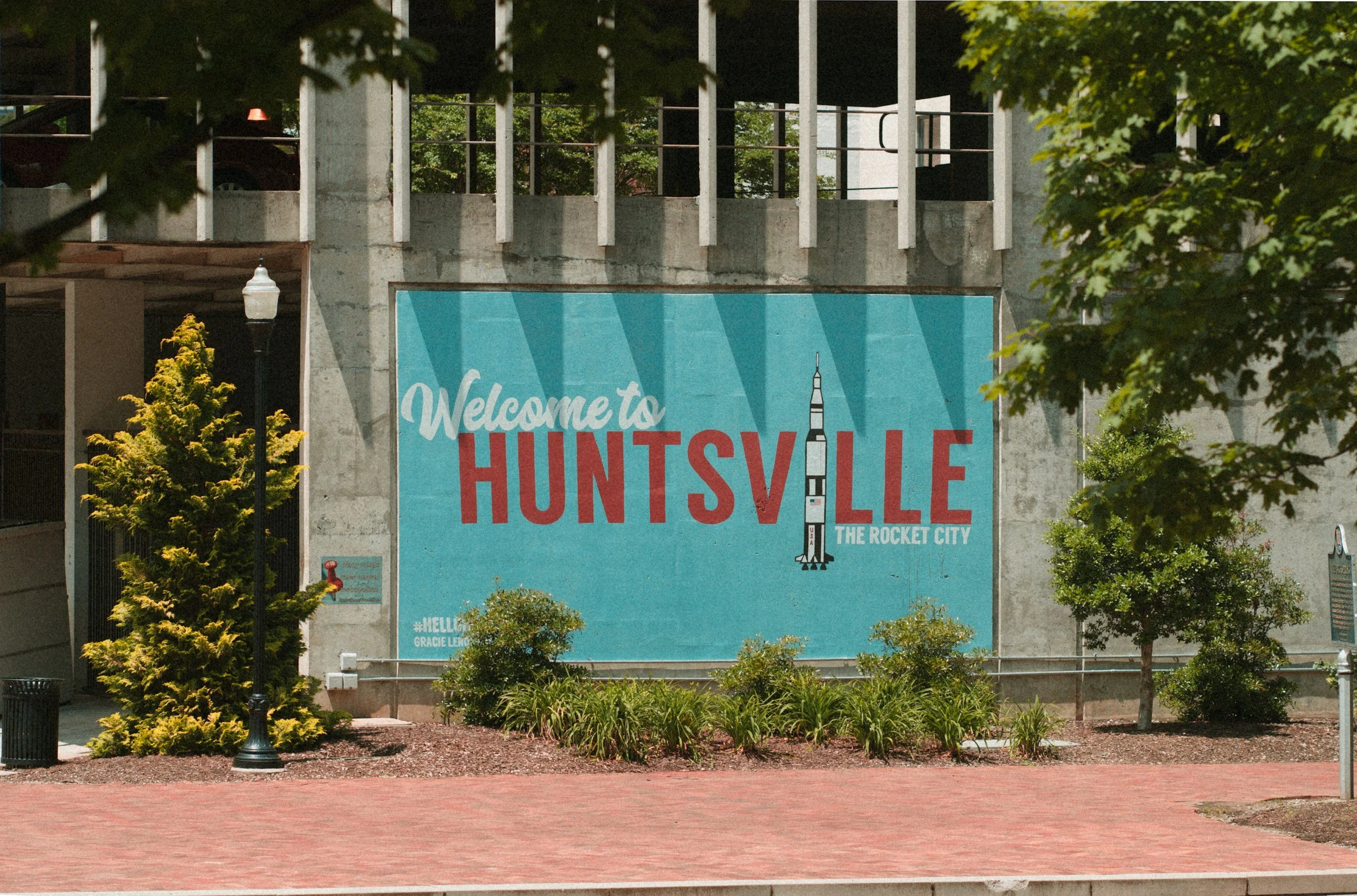 Large sign that reads "Welcome to Huntsville, The Rocket City" with an illustration of a rocket and a hashtag #HELLO, on a concrete wall surrounded by trees, lamp posts, and a park area.