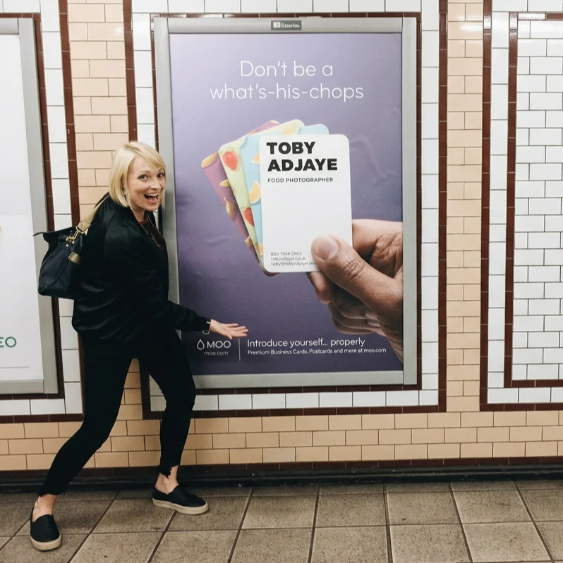 A woman with blonde hair smiling and pointing at a large advertisement in a subway station. The ad features business cards reading 'Toby Ajaye, Food Photographer' and a hand holding the cards. The background of the ad has the phrase "Don't be a what's-his-chops."