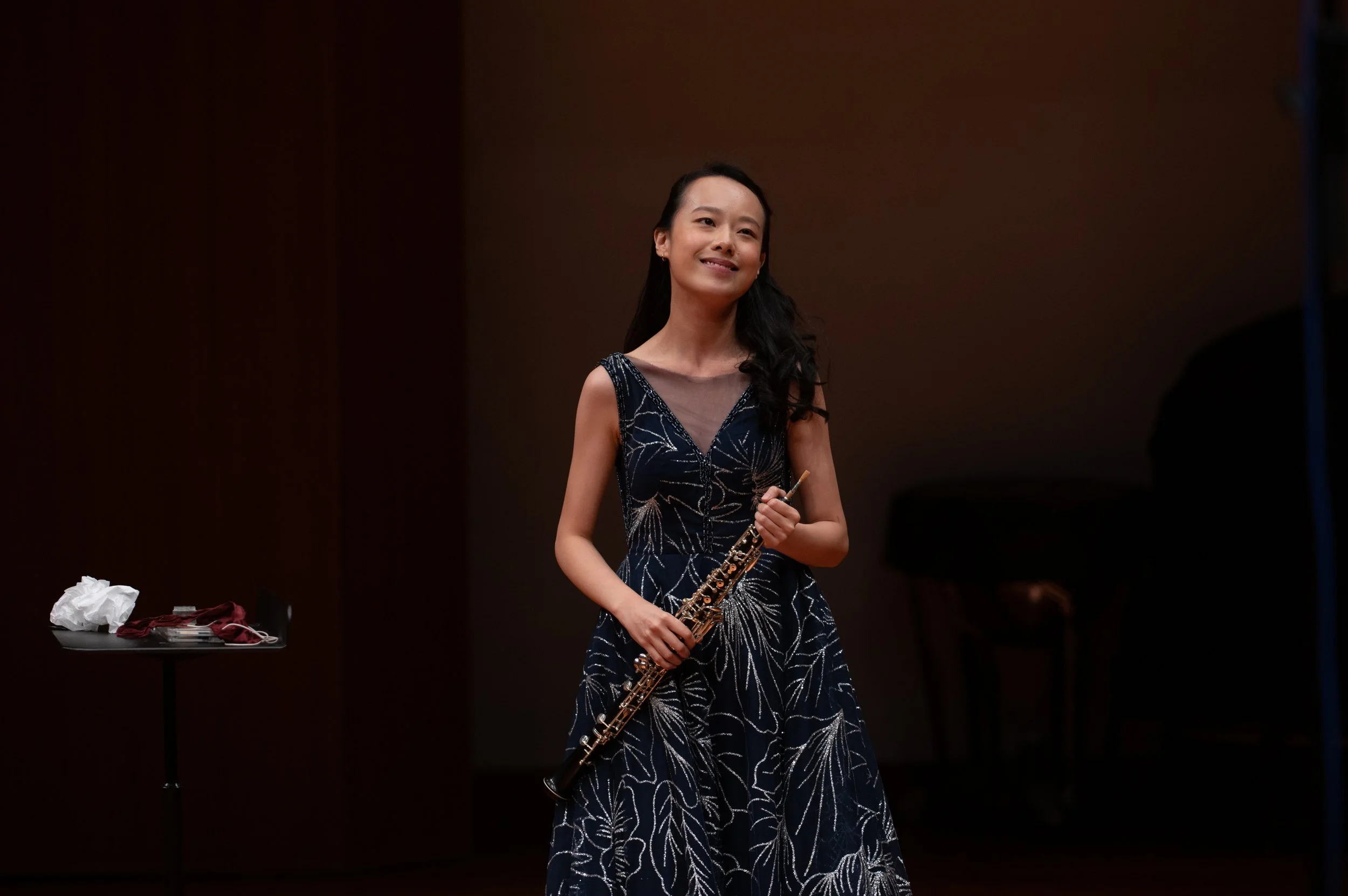 A woman in a dark dress with a floral pattern holding a musical instrument, standing on stage with a dark background.