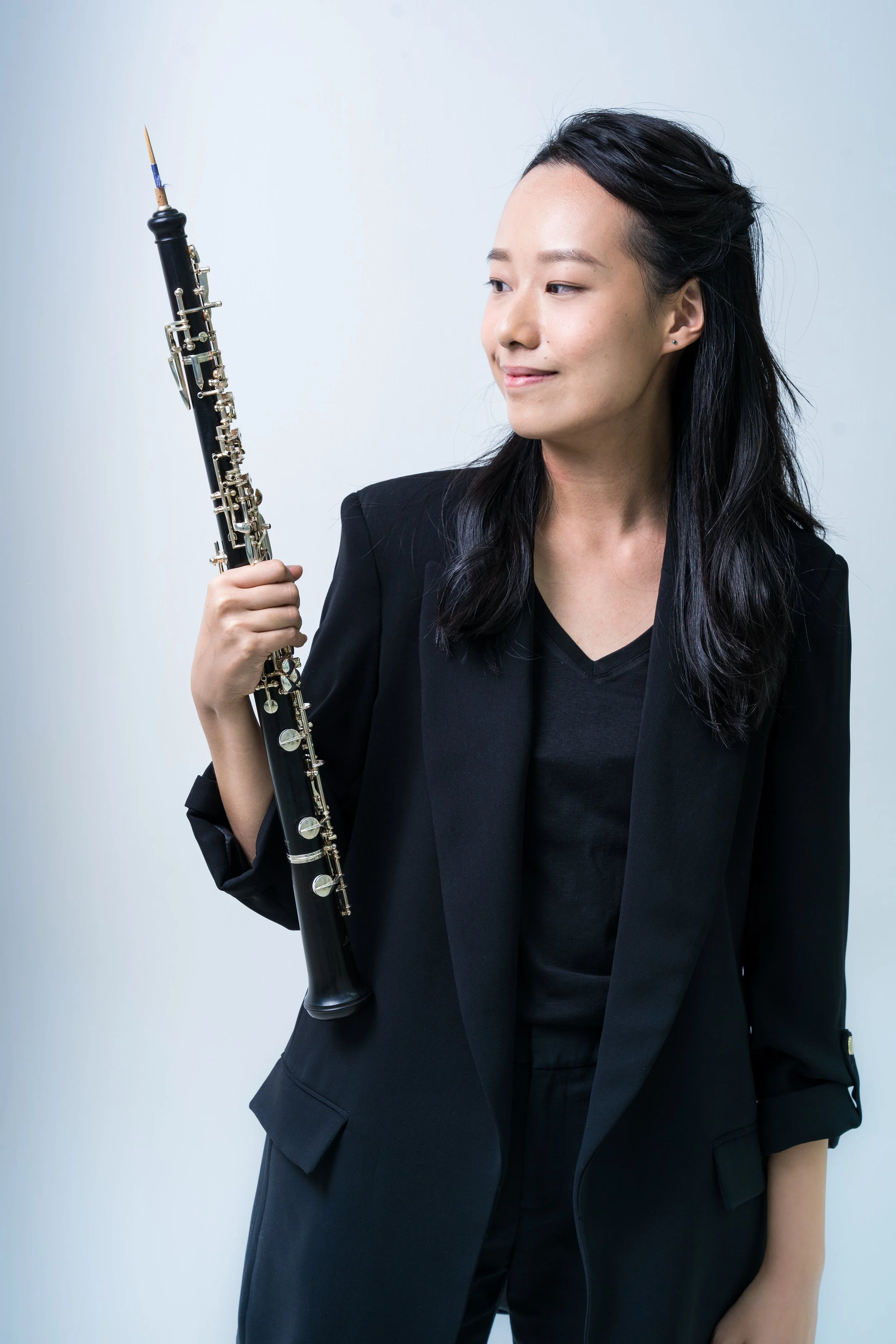 A woman in a black blazer holding a clarinet against a plain light background.