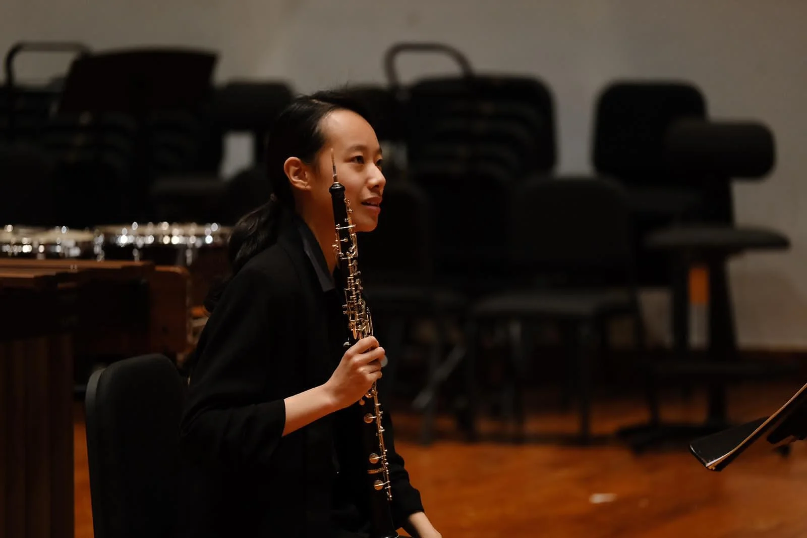 A woman sitting in a music room, holding a musical instrument (oboe), with stacked chairs and music stands in the background.