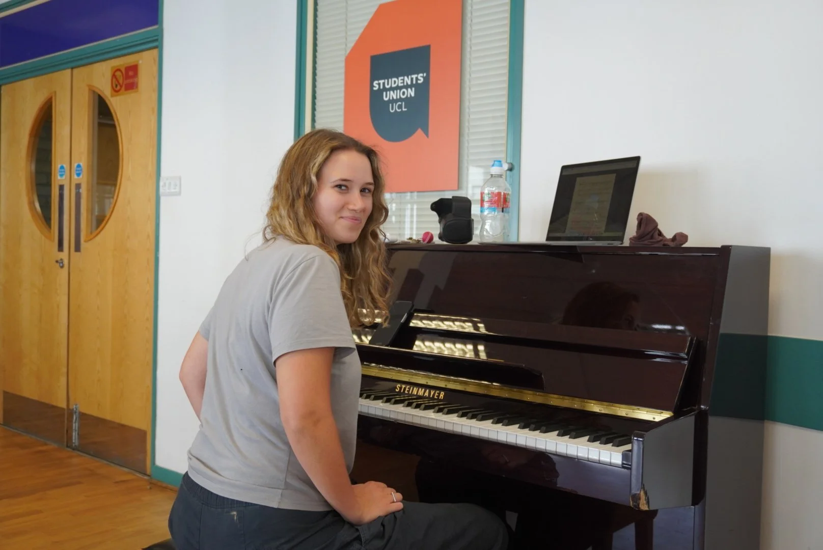 A young woman with curly brown hair sitting at a black Steinmayer upright piano in a room with wooden floors and walls painted white and green. She is smiling while looking back at the camera. There is a laptop, a water bottle, and other items on top of the piano. A sign on the wall behind her reads 'Students' Union UCL'.