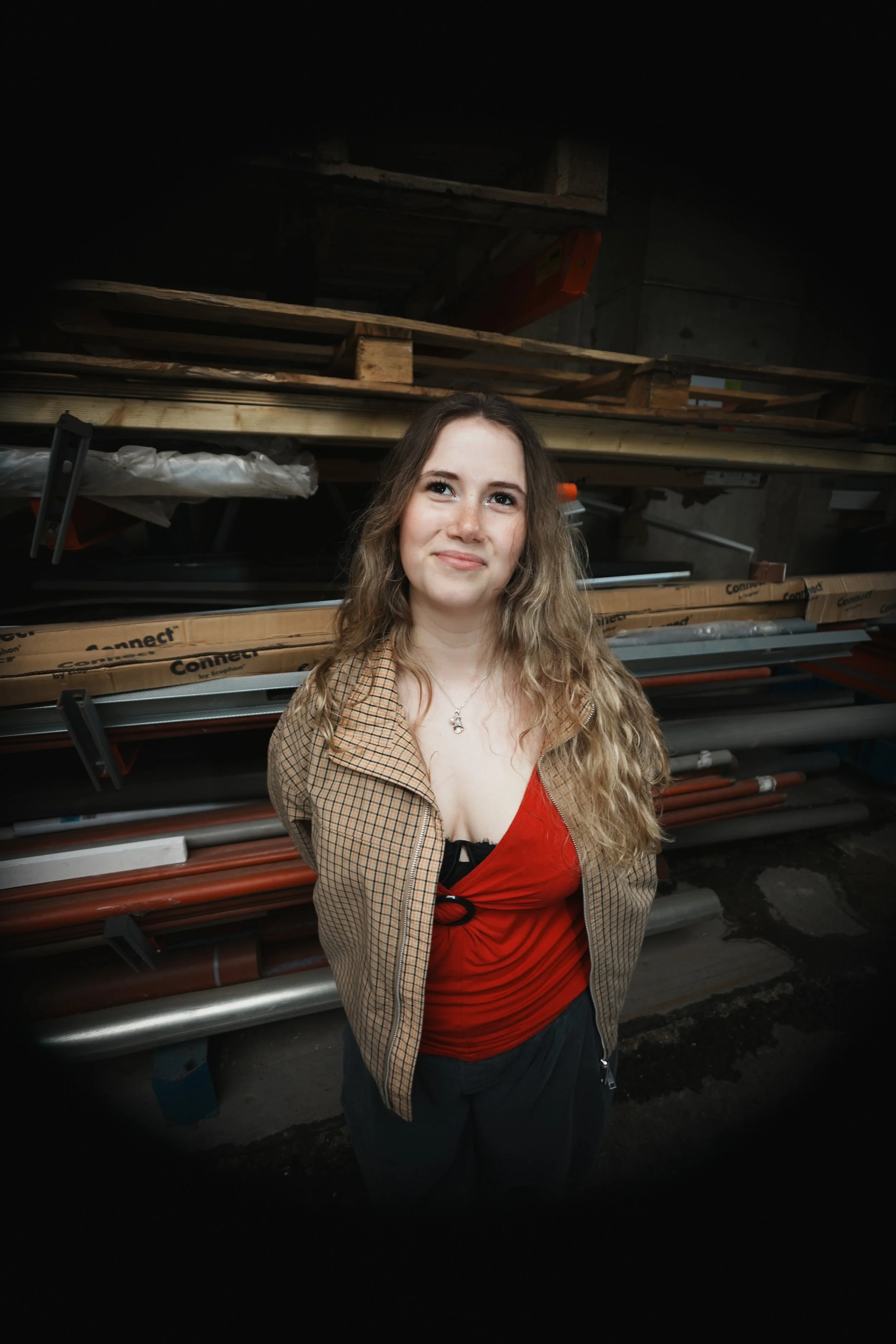A young woman with long wavy hair standing and smiling with wooden building supplies behind her.