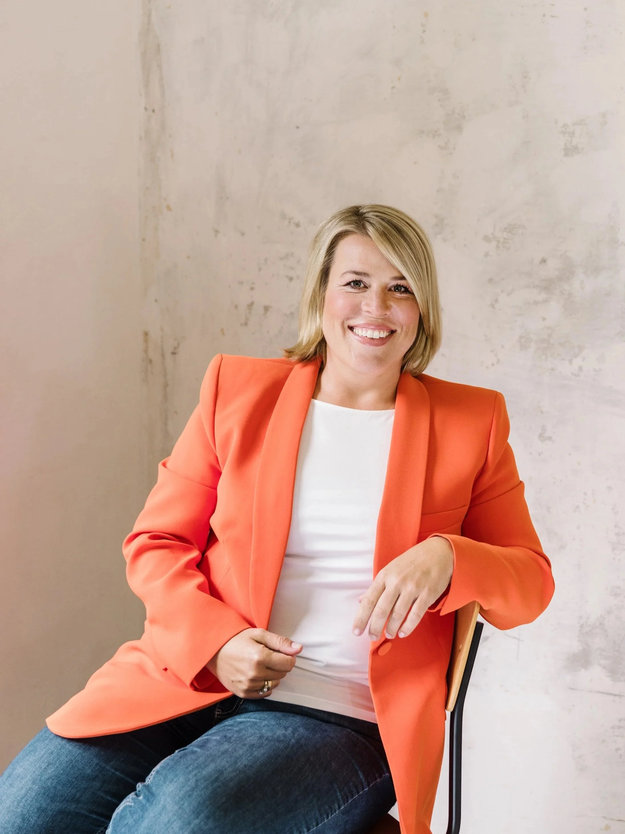 Frau mit blondem Haar, lächelnd, trägt eine orangejacke und eine weiße Bluse, sitzt auf einem Stuhl vor einer hellen Wand.