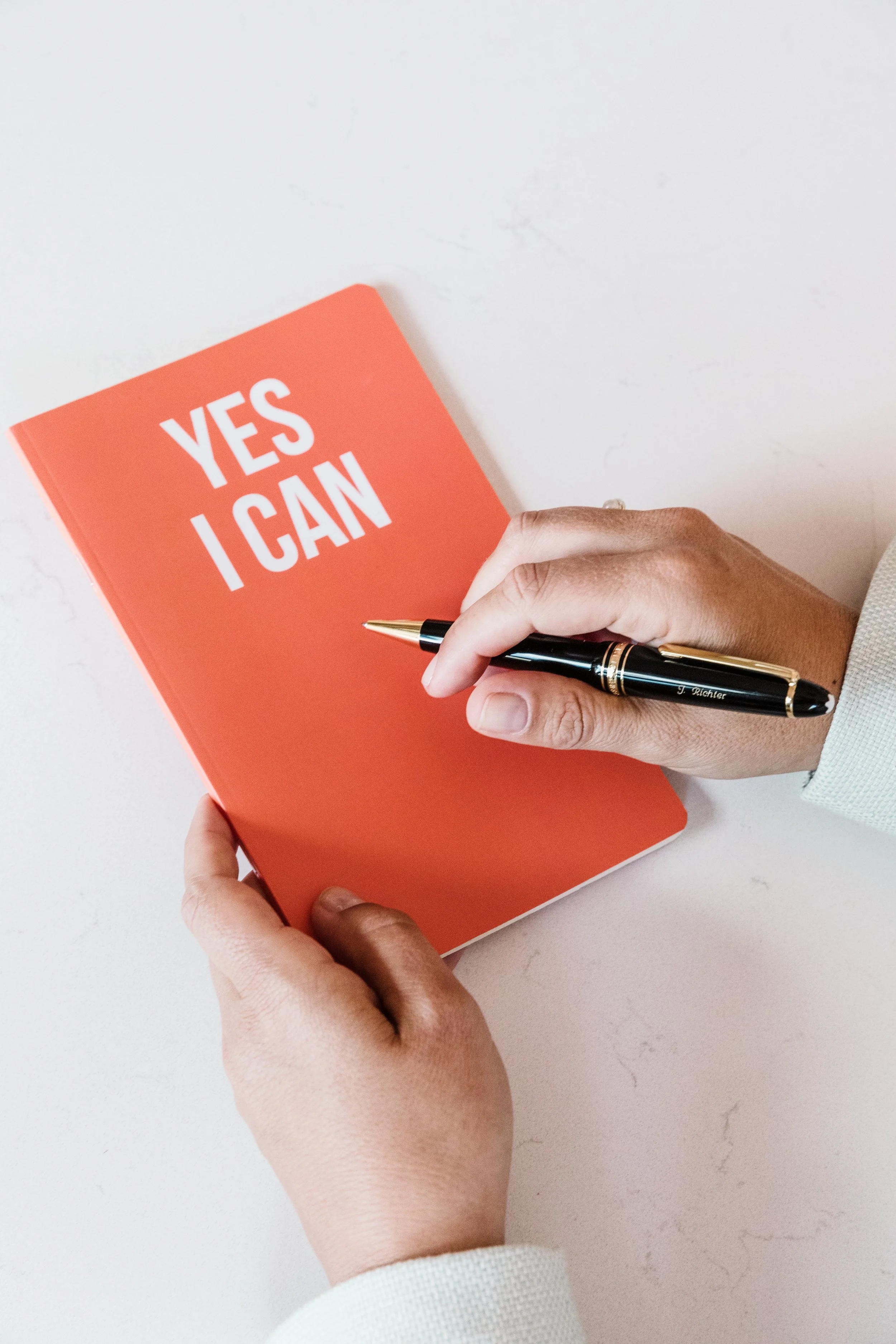 Eine Hand hält einen schwarzen Kugelschreiber vor einem orangefarbenen Notizbuch mit weißen Buchstaben, auf dem steht 'YES I CAN', auf einem hellen weißen Tisch.