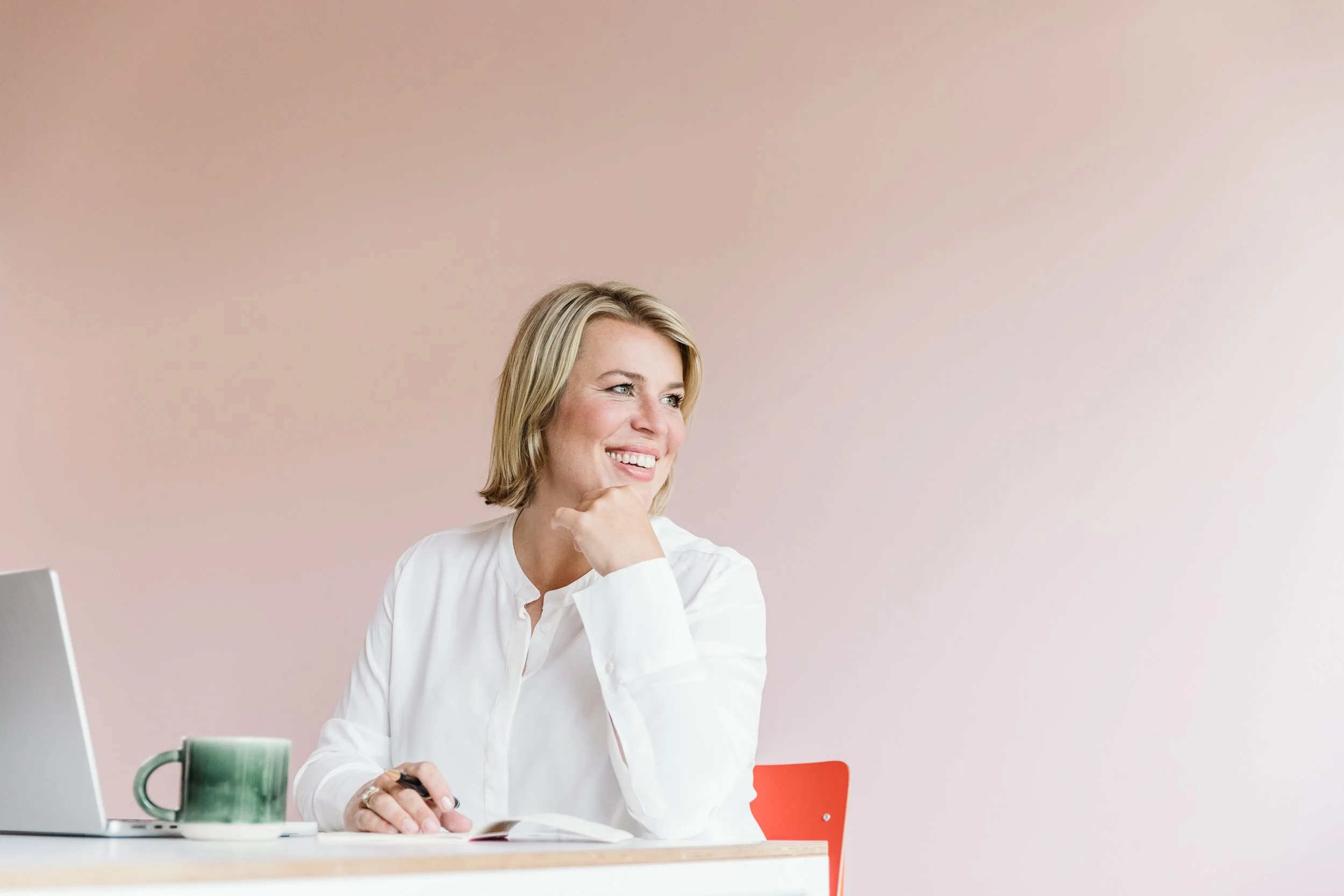 Eine lachende Frau mit blonden Haaren, die an einen Tisch sitzt, einen Kugelpen in der Hand hält, vor einem Laptop und einer Tasse. Im Hintergrund ist eine pinkfarbene Wand.