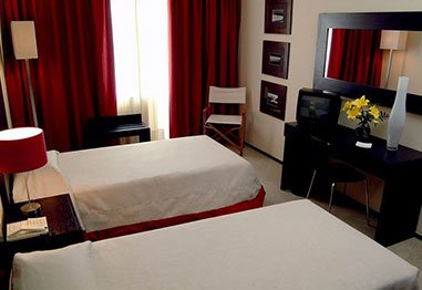 Modern hotel room with two beds, red curtains, black furniture, wall mirror, and yellow flowers.