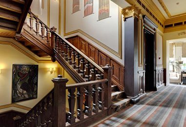 Elegant historic home interior with a grand wooden staircase, ornate railings, framed artwork, and patterned hallway carpet