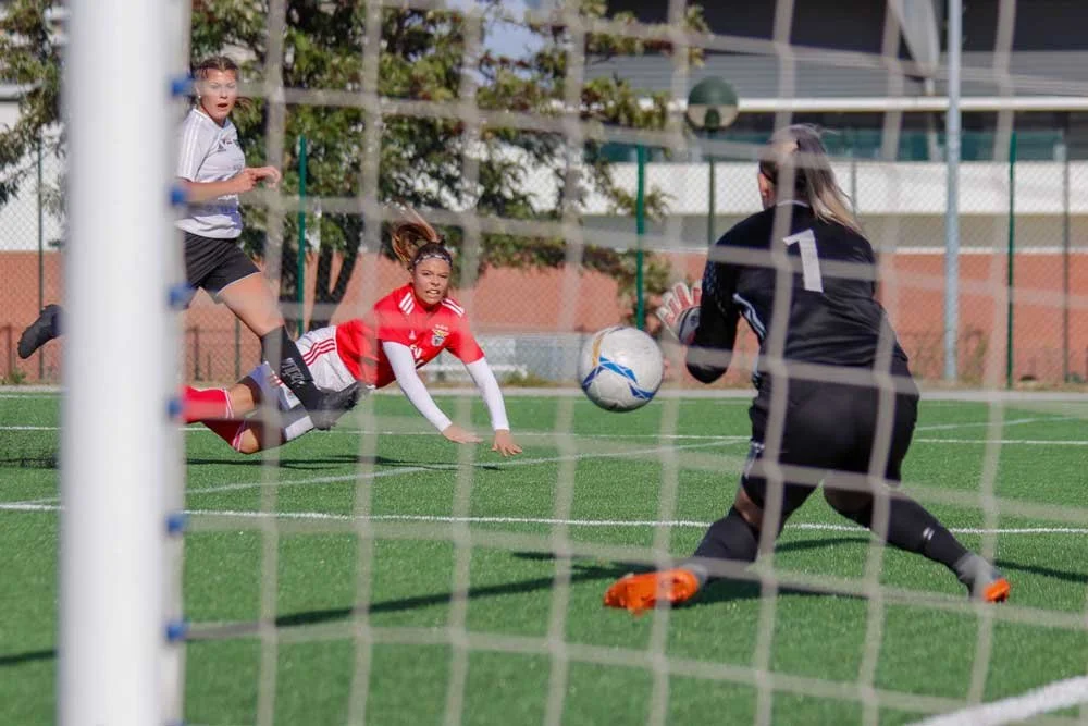 Women's soccer players compete near the goal as a goalkeeper in black dives toward the ball.