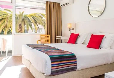 Bright modern bedroom with a white bed, red accent pillows, striped blanket, palm tree views, and round wall mirror