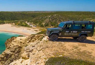 Black off-road tour vehicle parked on a rocky coastal cliff overlooking a turquoise cove and sandy beach